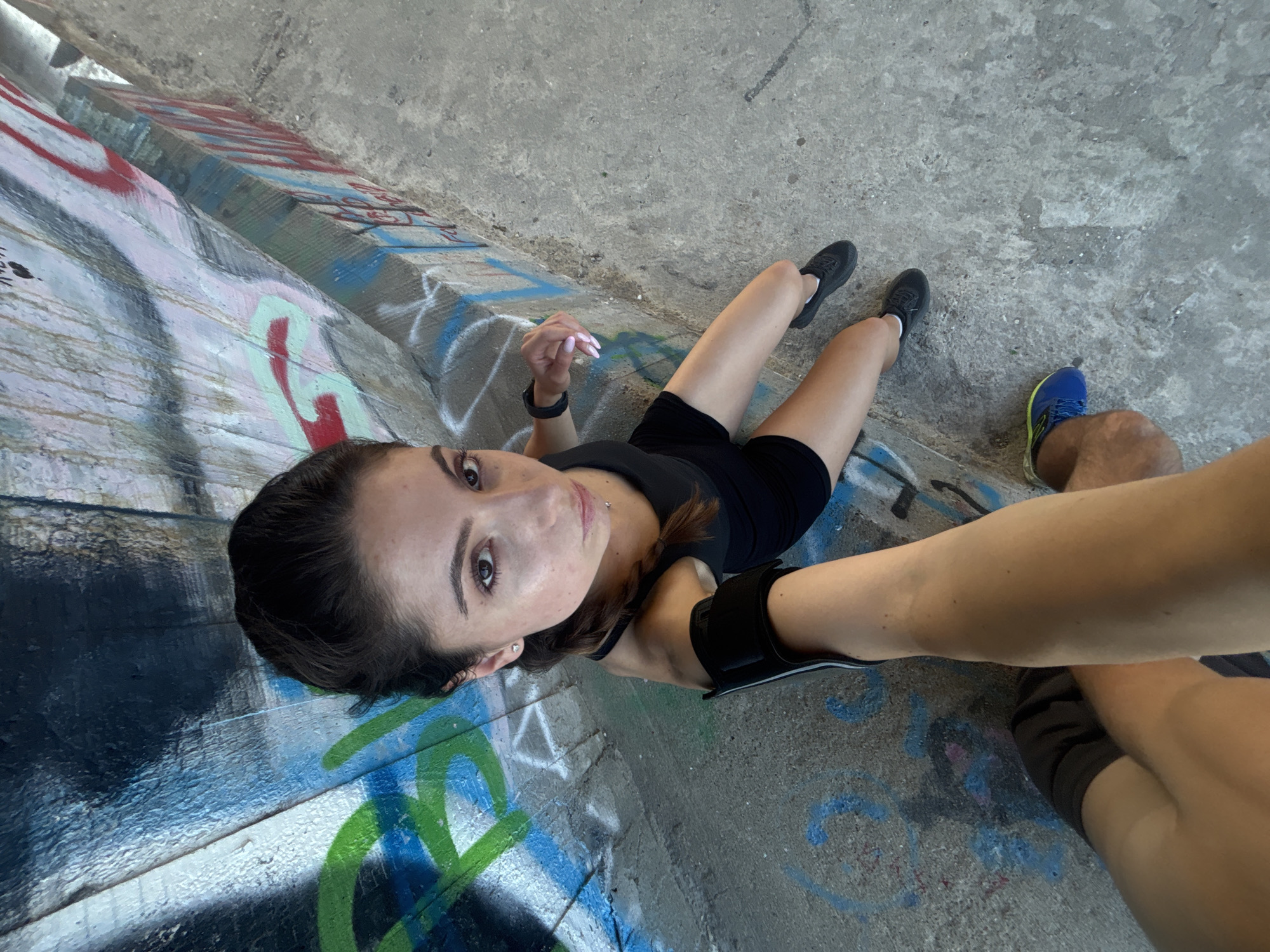 This media file is a vertically oriented, high-angle photograph taken outdoors. The scene depicts two individuals, one predominantly in the foreground and another partially visible in the background.
The primary subject is a young woman with dark hair, wearing a black tank top and black shorts. She has a black armband on her left arm and is looking directly at the camera. Her expression appears neutral. Her legs are extended, and she is wearing black athletic shoes.
The secondary subject is a person whose legs and feet are visible in the lower right portion of the frame. This individual is wearing grey shorts and blue and yellow athletic shoes.
They are positioned on a concrete surface with visible graffiti. The graffiti features various colors, including red, pink, light blue, green, and black. The concrete appears weathered and cracked in places, suggesting an urban or industrial setting. The background consists of more concrete structures, also marked with graffiti.
The lighting suggests it is daytime, with natural light illuminating the scene. The presence of graffiti and the concrete structures might indicate a location like an underpass, a skate park, or an abandoned industrial area. The overall mood is casual and perhaps a moment captured during an outdoor activity or break.
There is no visible text in the image. The crops provided focus on specific areas of the scene, highlighting the graffiti, the footwear of the individuals, and close-ups of the woman's face and arm. The location context provided is Varna, Bulgaria.