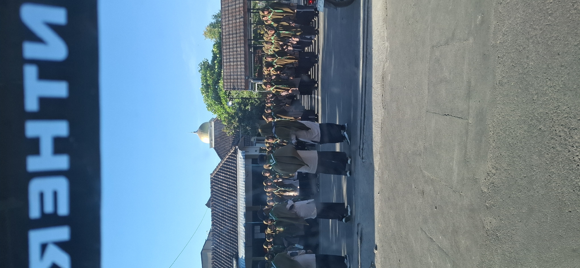 A group of individuals, likely students, are gathered in an outdoor setting, possibly a school yard or public space. They are dressed in uniforms that appear to be a light-colored shirt with dark pants or skirts, and many are wearing brown or olive-green cloaks or sashes. The people are standing in rows, suggesting a formation or a ceremony. In the background, there are buildings with tiled roofs, and a prominent golden dome is visible, which may indicate a religious structure such as a mosque or a church. Trees are also present, adding to the outdoor environment. The sky is clear and blue, suggesting a sunny day. The foreground shows a paved surface, possibly a road or courtyard, with some visible cracks and patches, indicating wear and tear. The overall scene appears to be a daytime event in a community.