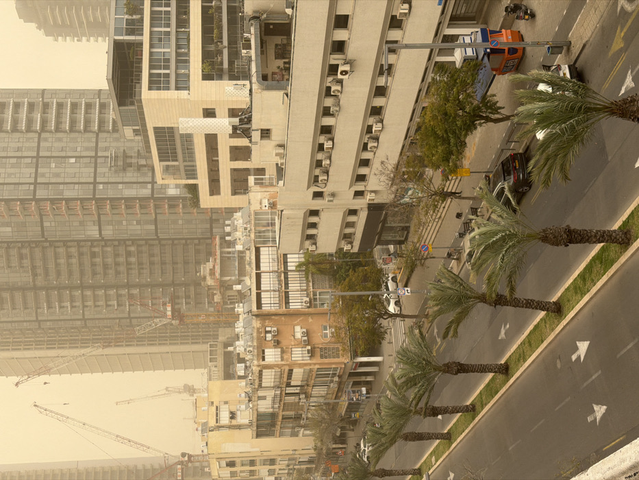 The image depicts an urban street scene in Tel-Aviv, Israel, under a pervasive, uniform yellowish-brown dust haze, significantly reducing visibility. The scene is observed from an elevated vantage point.
In the immediate foreground, an asphalt road features two visible lanes with white directional arrow markings. A narrow green median strip separates these lanes, hosting a continuous row of approximately seven date palm trees with textured trunks and green fronds, extending diagonally from the bottom-left.
To the right of the road, a sidewalk runs parallel, featuring parked vehicles, pedestrians, and street furniture. Parked vehicles include a dark-colored SUV and two white cars further down the street. Two scooters or motorcycles are parked near a distinct orange and blue street kiosk. A lone human figure, dressed in dark attire, walks on the sidewalk near a tree. A small dark-colored animal, possibly a dog, is visible near a street bench. Streetlights and traffic signs are also present along the sidewalk.
Buildings of varying architectural styles line both sides of the street. On the left, several older, multi-story residential or commercial buildings feature balconies, numerous externally mounted air conditioning units, and rooftop solar water heaters and satellite dishes. One building displays a "for rent" sign in Hebrew. On the right, a multi-story building with numerous horizontally aligned windows and many air conditioning units is visible. Adjacent to it stands a modern, light-colored building with large windows and terraced levels, topped by a tall, narrow, white cylindrical structure.
In the midground and background, the Tel-Aviv skyline is dominated by numerous high-rise buildings, many of which appear to be under active construction. Multiple red and white lattice boom tower cranes are visible, reaching above the partially constructed and completed skyscrapers. The pervasive dust haze heavily obscures the upper sections of these buildings and the distant horizon, creating a muted, sepia-toned urban landscape.