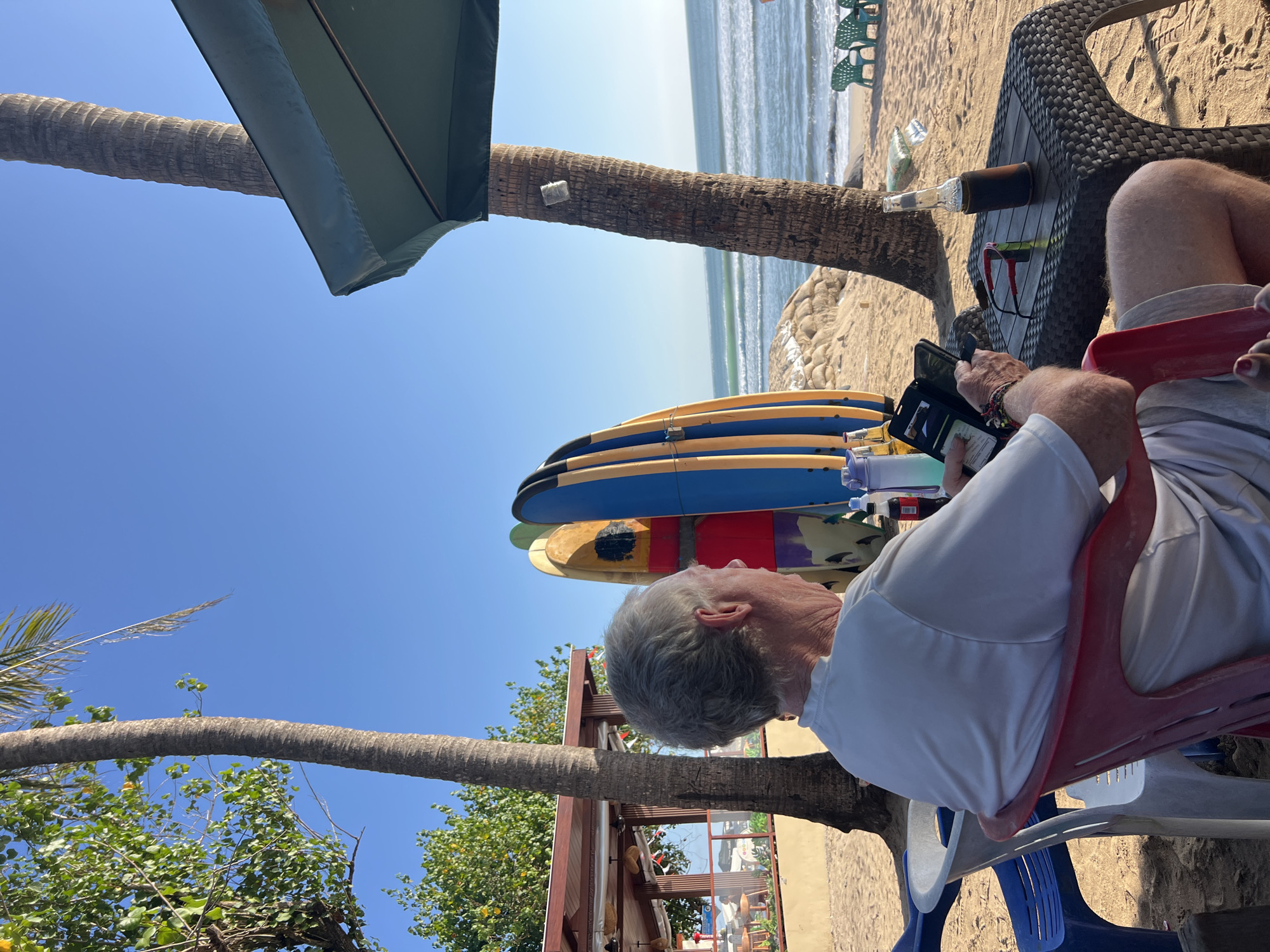 An elderly Caucasian male with gray hair is sitting in a red plastic chair on a sandy beach in Kuta, Indonesia. He is wearing a white t-shirt and light-colored shorts, and is looking at an open black wallet with a phone in it, which he is holding in his hands. He is seated at a dark woven plastic table with a bottle of dark liquid in a holder, a pair of red-framed sunglasses, and a bottle of water. Behind him, several surfboards are propped upright. Palm trees are visible, and in the background, the ocean with gentle waves and a clear blue sky can be seen. A green umbrella is partially visible in the upper right corner. The scene appears to be a relaxed beachfront setting.
