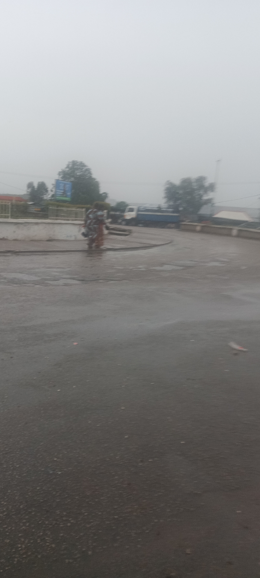 The image shows a street scene in Kuru Dungus, Nigeria on a heavily overcast day. The ground is wet, presumably from rain. A person is walking along a sidewalk carrying items on their head. A truck is parked on the side of the road near a white wall and railing that separates the street from another area, possibly a drop-off or retaining wall. In the background are trees, power lines, and a billboard.