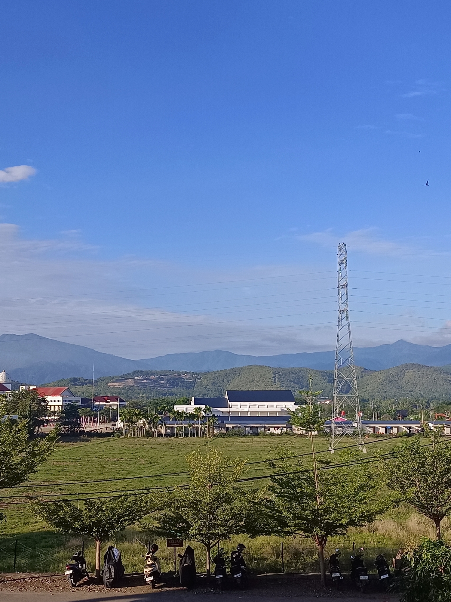 The image depicts an outdoor scene with a wide expanse of clear blue sky, featuring a few wispy white clouds and two birds in flight. Below the sky, a mountain range forms the backdrop, with layers of green hills and more distant, hazy mountains. In the middle ground, there are several buildings, including one with a white dome and red roof, and a larger, modern building with a dark roof. A prominent metal pylon with electrical wires stretches vertically through the scene. A large, grassy field occupies a significant portion of the middle ground, with rows of trees lining the foreground. Several motorcycles are parked along a dirt path in the immediate foreground, some covered with black tarps, and one uncovered with a rider. A sign that reads "KELENG" is visible on a post near the trees. The overall impression is a view of a semi-rural or suburban area with natural landscapes and infrastructure, likely situated in Manyang Cut, Indonesia, given the provided context.