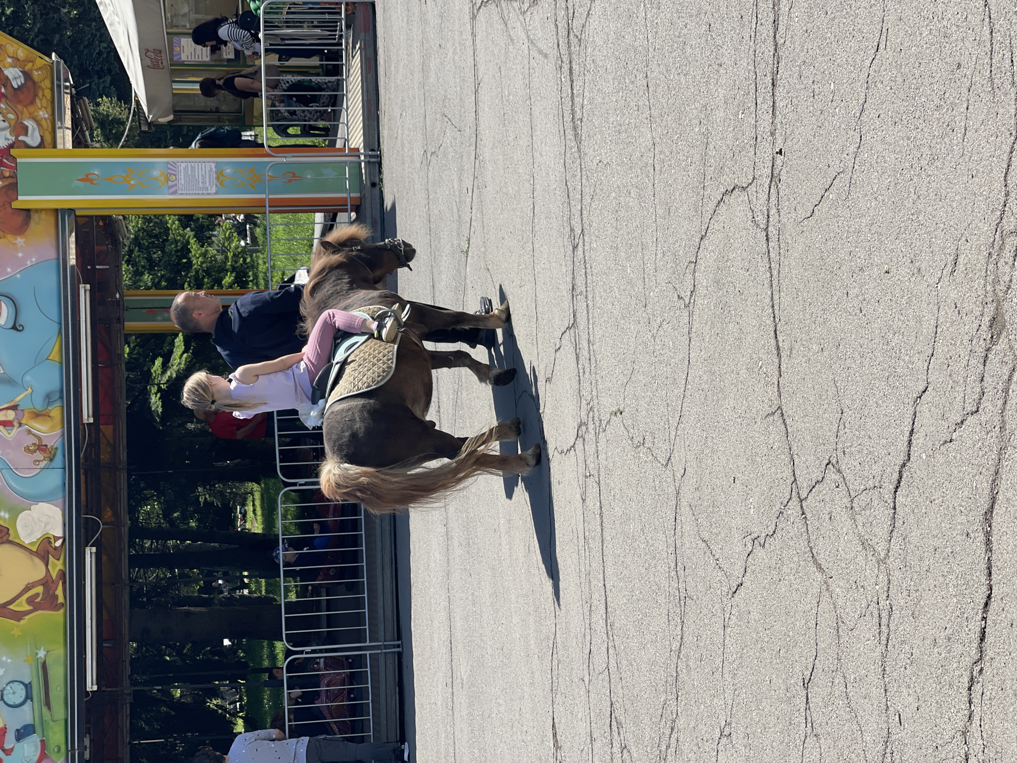 A photograph captures a moment at an amusement park, possibly in Sofia, Bulgaria, given the Eastern European context. A young, fair-haired girl rides a small pony. A man walks alongside the pony, holding onto the reins, presumably to guide it. The pony is equipped with a saddle. The surroundings suggest a bustling amusement park. An elaborate carousel structure can be seen in the background. A Coca-Cola-branded awning is also visible. The ground is paved and displays cracks in the surface. Additional blurred figures are present, indicating the presence of other park-goers. The scene seems to be a snapshot of leisure activities in a public space.
