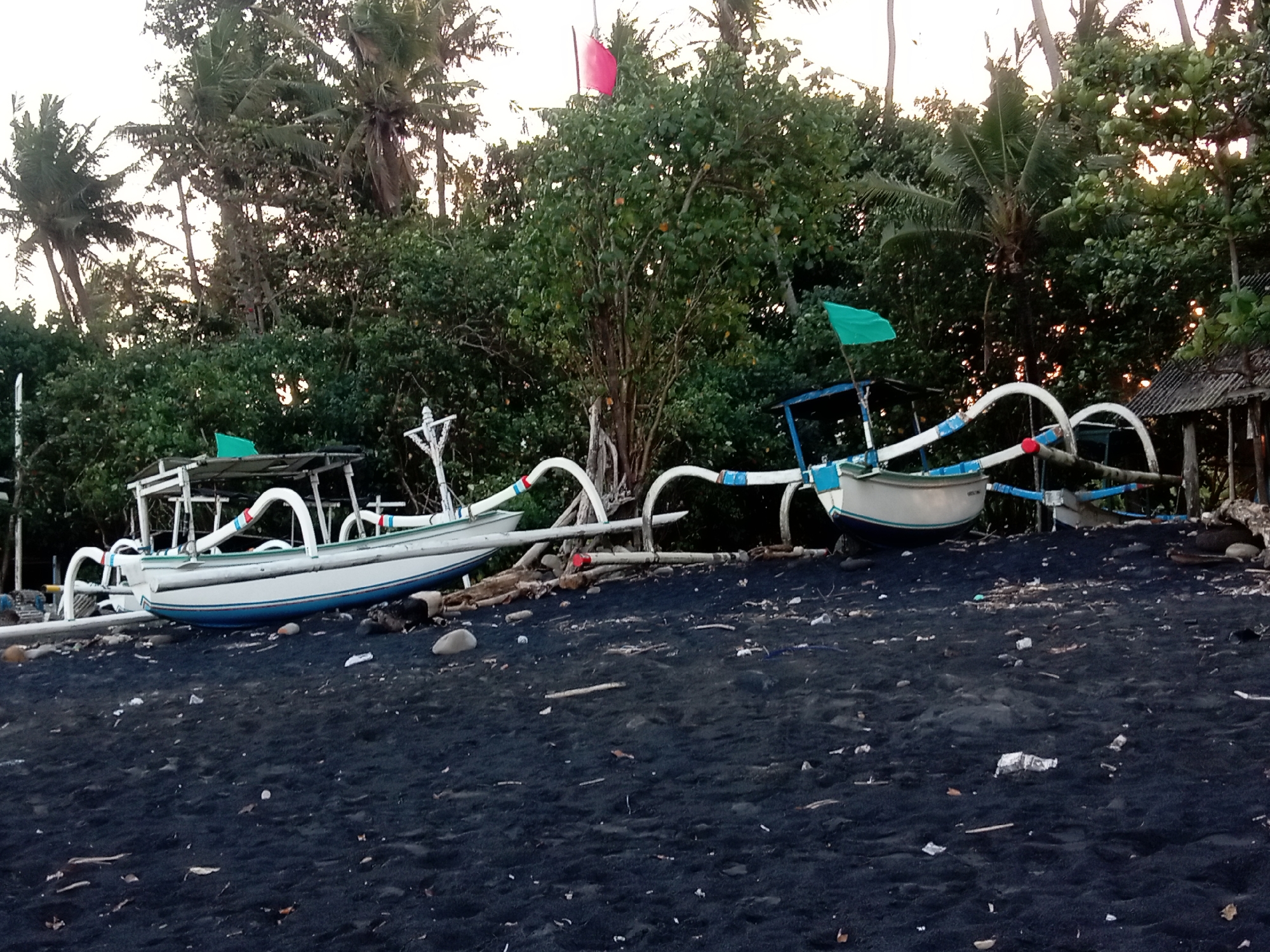 This media file shows two traditional Balinese fishing boats resting on a black sand beach. The boats are white with blue stripes and feature distinctive curved outriggers. One boat is closer to the foreground and the other is further back. A green flag flies from a pole on each boat. In the background, there is lush green foliage, including palm trees, and a small, rustic structure with a corrugated roof. The sky is overcast, suggesting it might be early morning or late afternoon. The beach is covered in dark sand with a scattering of debris, including small rocks, driftwood, and some litter. The overall scene evokes a sense of tranquility and stillness, typical of a coastal setting in Bali. There is no visible text in the image.