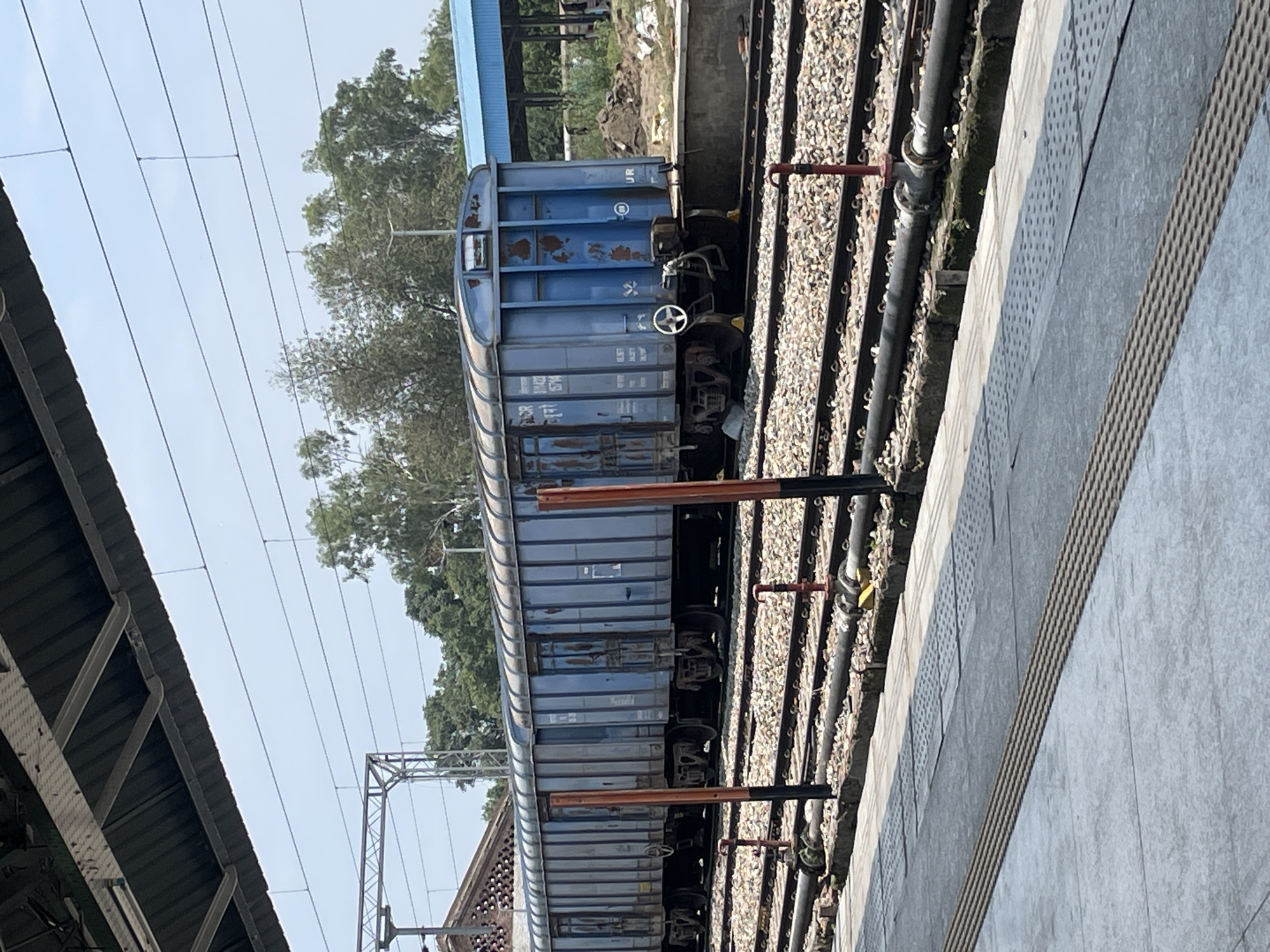 The image shows a blue train car parked on railway tracks at a station in Pathankot, India. The train car is a cargo wagon with multiple doors and visible wear and tear, including rust spots. It is situated alongside a concrete platform with tactile paving for visually impaired individuals. In the background, there are trees, power lines, and a blue-roofed structure, possibly a shelter or building at the station. The scene appears to be under a bright, clear sky, suggesting daytime. There are no people visible in the image.