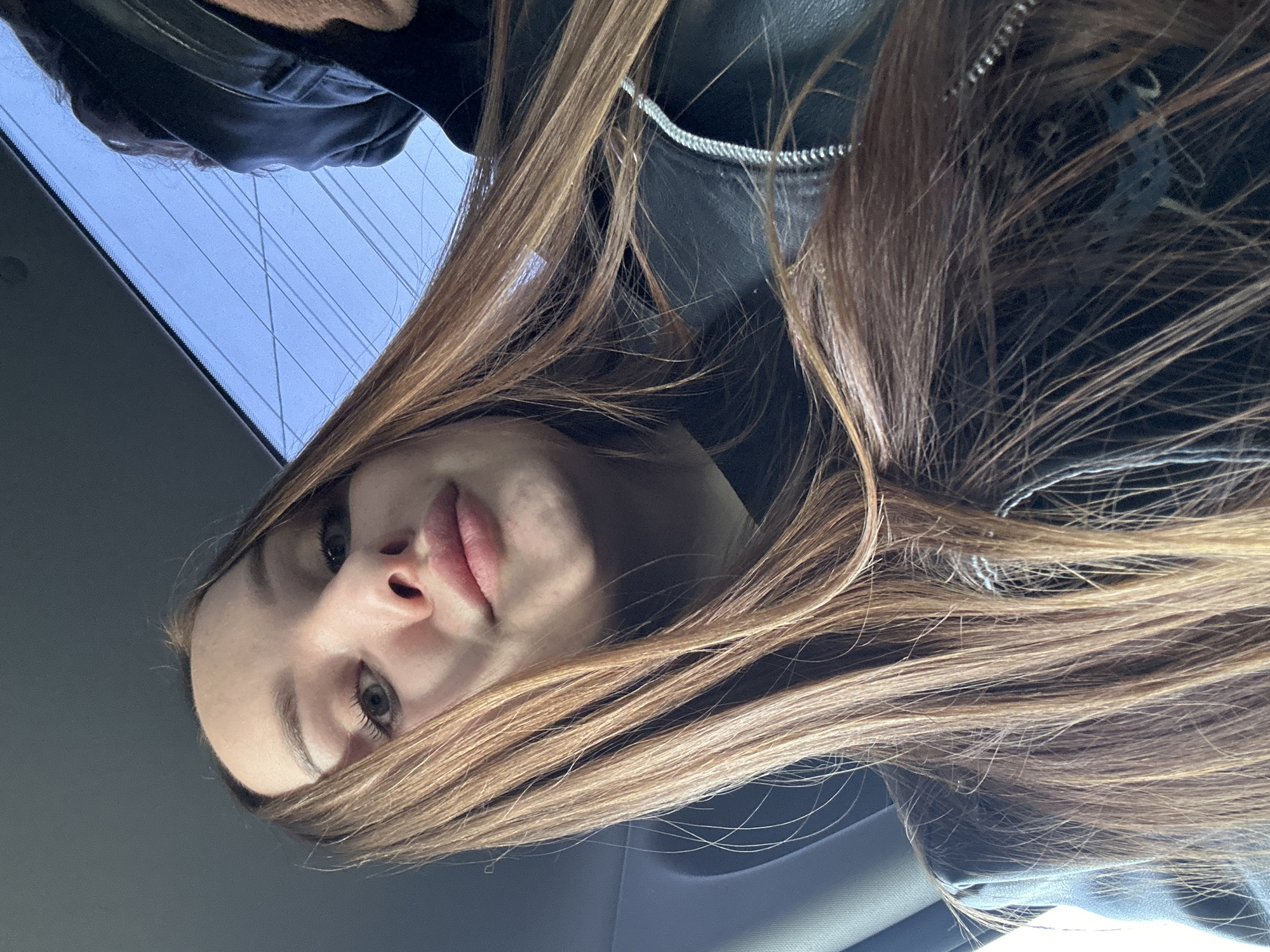 The image is a selfie taken from a low angle, looking up at a young woman with long, light brown hair. She has fair skin, prominent lips, and visible eyes. She is wearing a black leather jacket. The background shows a view of the sky through a car window, suggesting she is inside a vehicle. The exact location within Chaikovsky, Russia is not identifiable from the image.