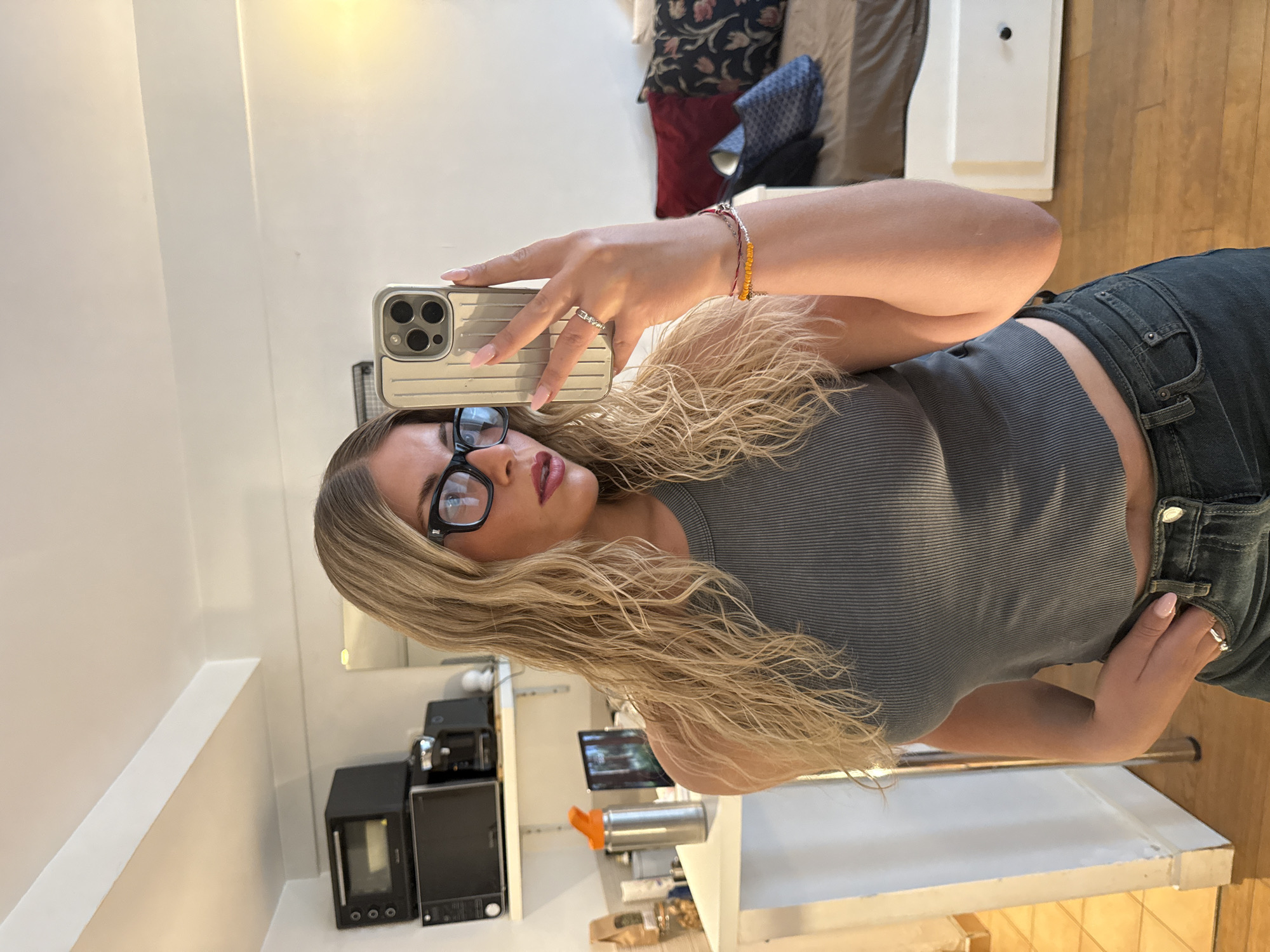 A blonde woman wearing glasses, a grey ribbed tank top, and dark jeans takes a mirror selfie. She is holding a silver iPhone in her right hand, which is raised to chest level. She is wearing rings on her left ring finger and right pinky finger, and bracelets on her right wrist. Her hair is long and wavy, and her nails are painted light pink. Behind her, there is a shelf with kitchen appliances, a water bottle, and other items. A bed with pillows is visible in the background, as well as a patterned fabric draped over the edge of the bed. The floor is hardwood.