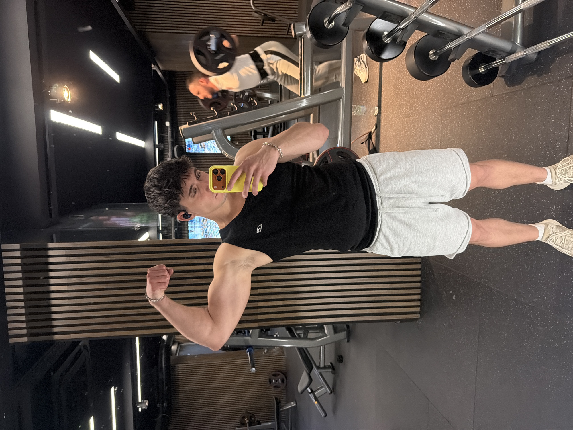 The image shows a young man posing in a gym, likely taking a selfie. He is wearing a black tank top and light gray shorts, with white sneakers. He's flexing his right bicep while holding a yellow smartphone in his left hand. A silver bracelet is visible on his left wrist. He has short dark hair and appears to have wireless earbuds in his ears.
The background reveals a well-equipped gym with weightlifting machines and a mirrored wall showing another person lifting weights. The gym has dark flooring and slatted wooden wall panels. Given the style of the gym equipment and the presence of the slatted wooden wall panels, the location is likely a modern gym in an urban setting. The specific gym location in Sofia, Bulgaria cannot be determined from the image alone.