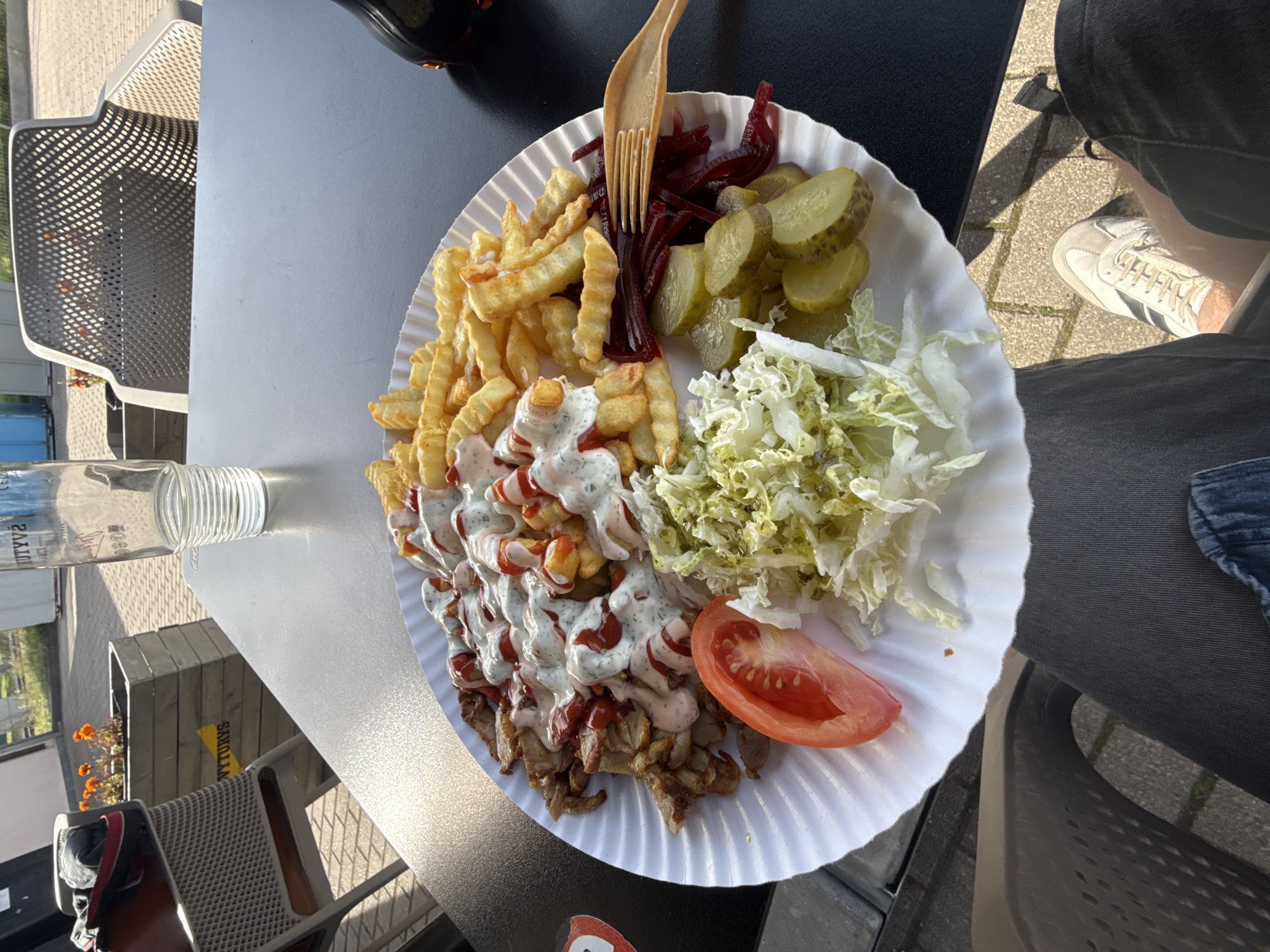 An outdoor scene at a casual dining establishment, likely in Šiauliai, Lithuania, shows a plate of food on a black table. The plate, made of white paper, contains a generous portion of shawarma meat, french fries, shredded lettuce, pickles, and a slice of tomato. Sauces, appearing to be a creamy white dressing and ketchup, are drizzled over the shawarma and fries. A wooden fork rests on the plate, near the pickles. In the background, a glass of water sits on the table, and a chair with a distinctive mesh design is visible. Further back, there are planters with orange flowers and a sign with yellow lettering on a wooden structure. The ground is paved with bricks. The image captures a sunny day, with shadows indicating the time of day. The perspective is from someone sitting at the table, with their legs and shoes in the foreground.