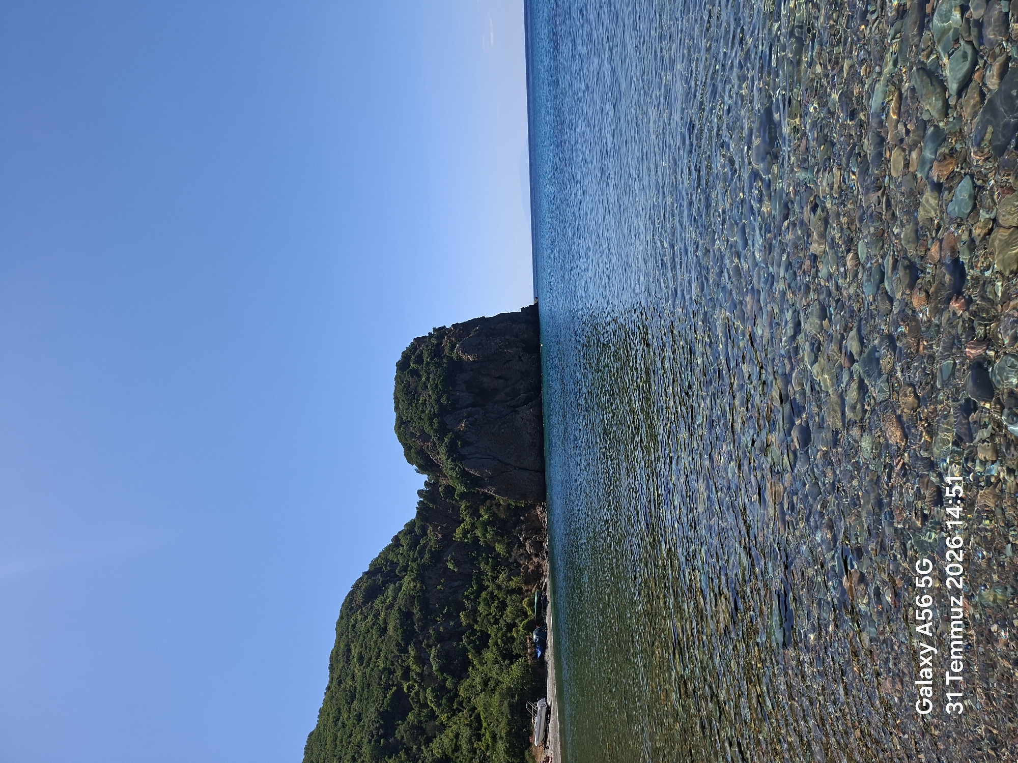 A bright, clear sky stretches over a tranquil seascape. In the foreground, shallow, crystal-clear water reveals a pebble-strewn seabed. Gentle ripples distort the view of the submerged stones.
Further out, the water deepens to a rich blue. A small cove is visible on the left, bordered by a lush, green, tree-covered hillside. A rocky outcrop, also covered in greenery, rises dramatically from the sea to the right of the cove.
On the shore of the cove, a white boat is beached, alongside what appears to be a blue tarp or cover and a dark green object, possibly another boat or equipment. The overall scene suggests a secluded and peaceful coastal location. The timestamp in the lower left corner indicates the image was captured in Gelibolu, Turkey on July 31, 2026, at 14:51.