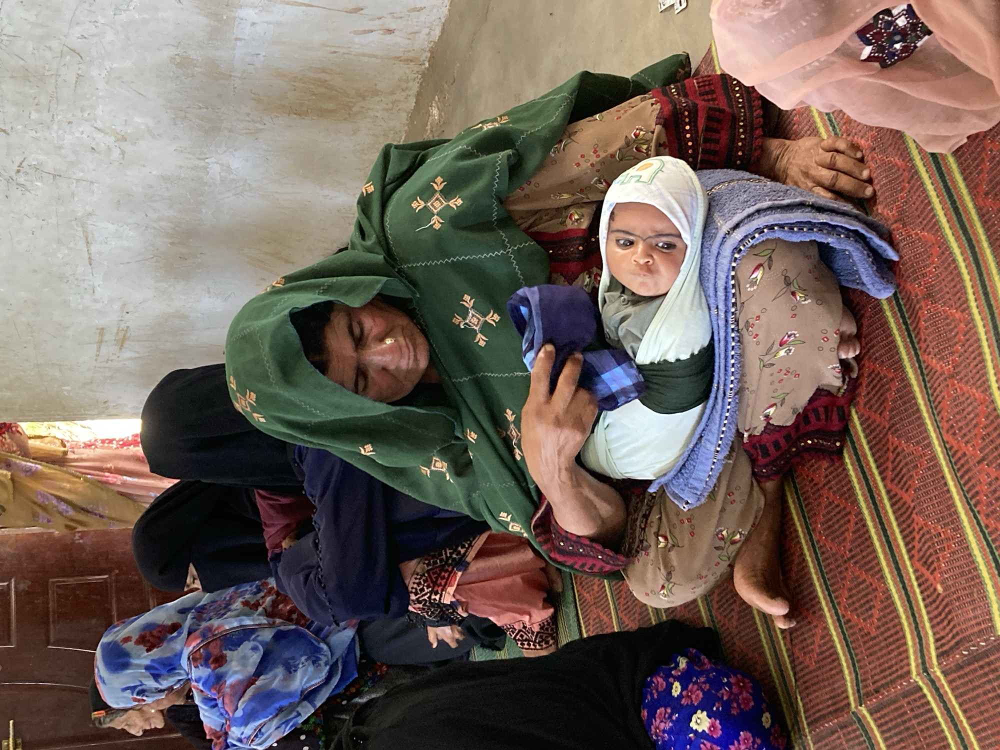 The photograph was taken indoors in the city of Goth Ali Marād, Pakistan. A group of women are sitting on a striped rug on the floor, interacting with a baby. One woman is holding the baby, who is wrapped in a white blanket with a blue blanket over it. The woman holding the baby is wearing a green headscarf. Other women are wearing various colored headscarves and garments. The room has a plain wall in the background. A partially visible dark wooden door is in the background, adorned with a golden decorative piece. The lighting appears to be natural.