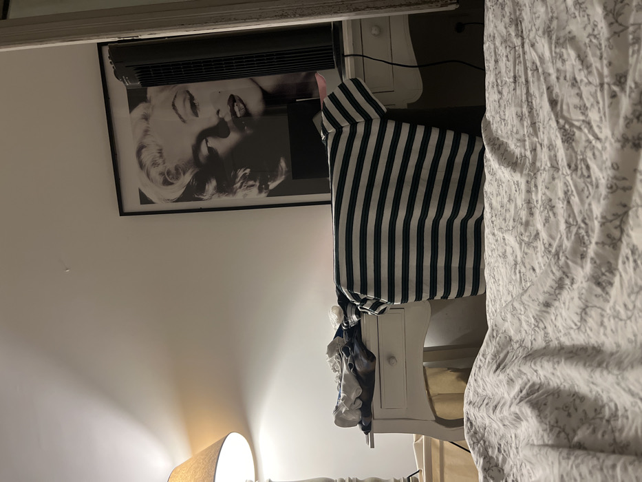 A bedroom features a white nightstand with a striped shirt hanging from its side. On the wall, there's a black and white framed picture of Marilyn Monroe and an air conditioning unit above it. To the left, a lamp emits a warm glow. A white bedspread with a delicate pattern dominates the right side of the frame.