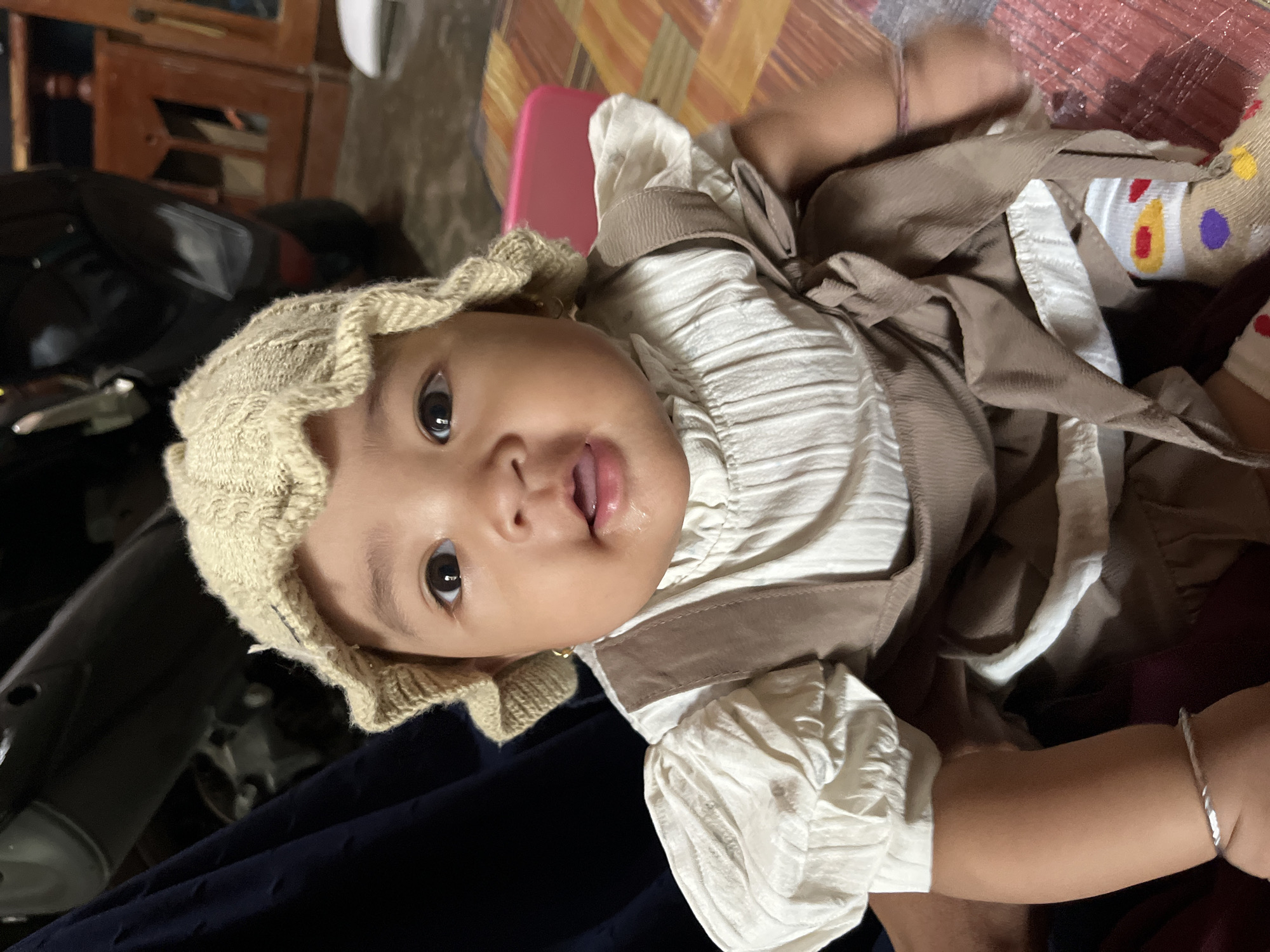 A baby girl is pictured from the chest up, looking directly at the camera. She is wearing a light beige knitted hat and a cream-colored shirt with puffed sleeves. Over the shirt, she wears a brown pinafore dress. Her mouth is slightly open, and she has a bit of drool on her chin. She has small gold earrings. Her left arm is visible, adorned with a silver bracelet. The background shows a dark blue cloth on the left, a motorcycle on the upper left, and a wooden cabinet on the right. The floor is covered with a mat with a colorful patterned design. The lighting suggests an indoor setting. The location is identified as Kruengmate, Indonesia.