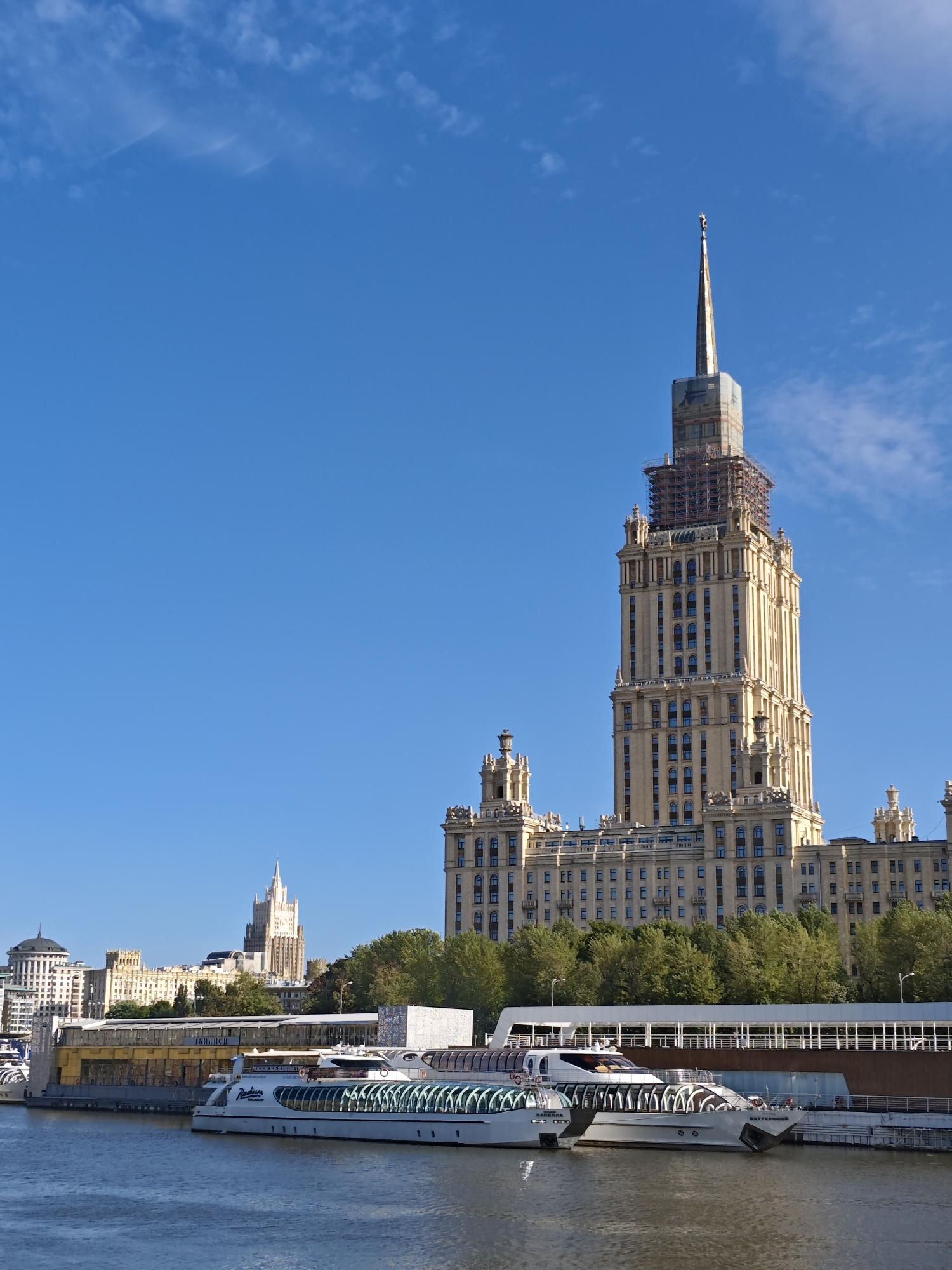 This is a vertical, medium-shot, eye-level, outdoor photograph taken on a clear, sunny day in Moscow, Russia. The photograph captures a wide view of the Moskva River with a prominent Stalinist skyscraper on the right and other buildings and boats in the foreground and background. The skyscraper, likely one of the Seven Sisters, features a golden facade, a tall spire, and scaffolding around its upper section, indicating ongoing construction or renovation. In the mid-ground, a long, modern building with large windows lines the riverbank, with trees behind it. Two white river cruise boats are docked in front of this building, one with "Radisson" visible on its side. Further in the background, other Stalinist-style buildings with spires are visible, contributing to the iconic Moscow skyline. The sky is a vibrant, cloudless blue.