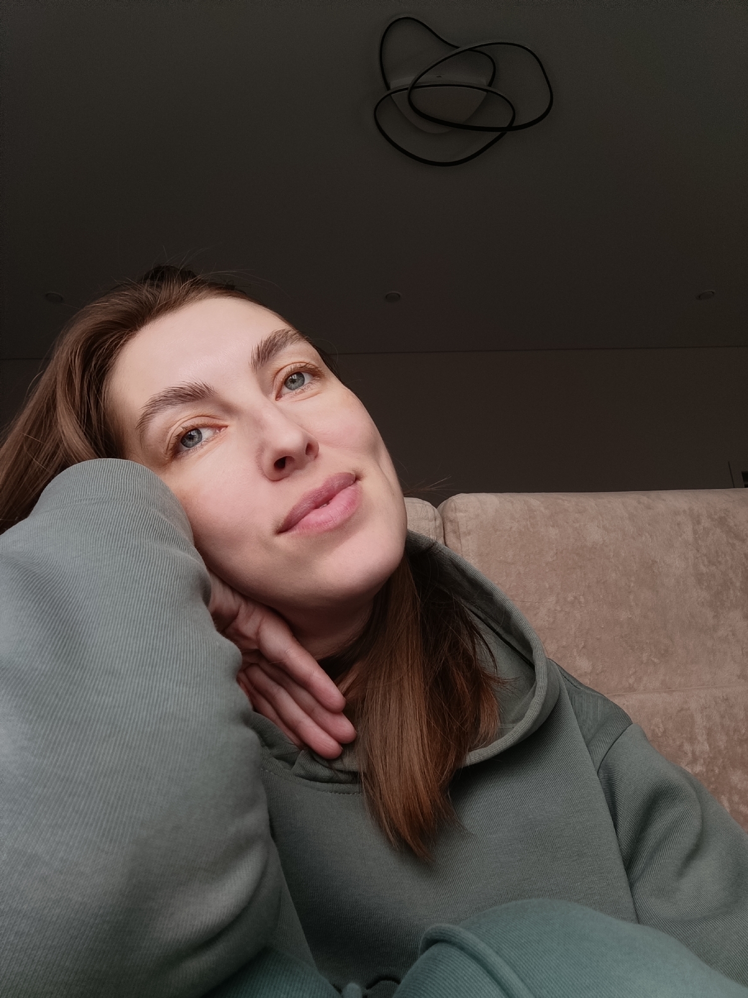 A young woman with light skin, blue eyes, and brown hair rests her head on her hand, wearing a green hooded sweatshirt. She is indoors. The background features a sofa or couch and a ceiling with a modern light fixture. The image was likely taken in an interior space in Chaikovsky, Russia.