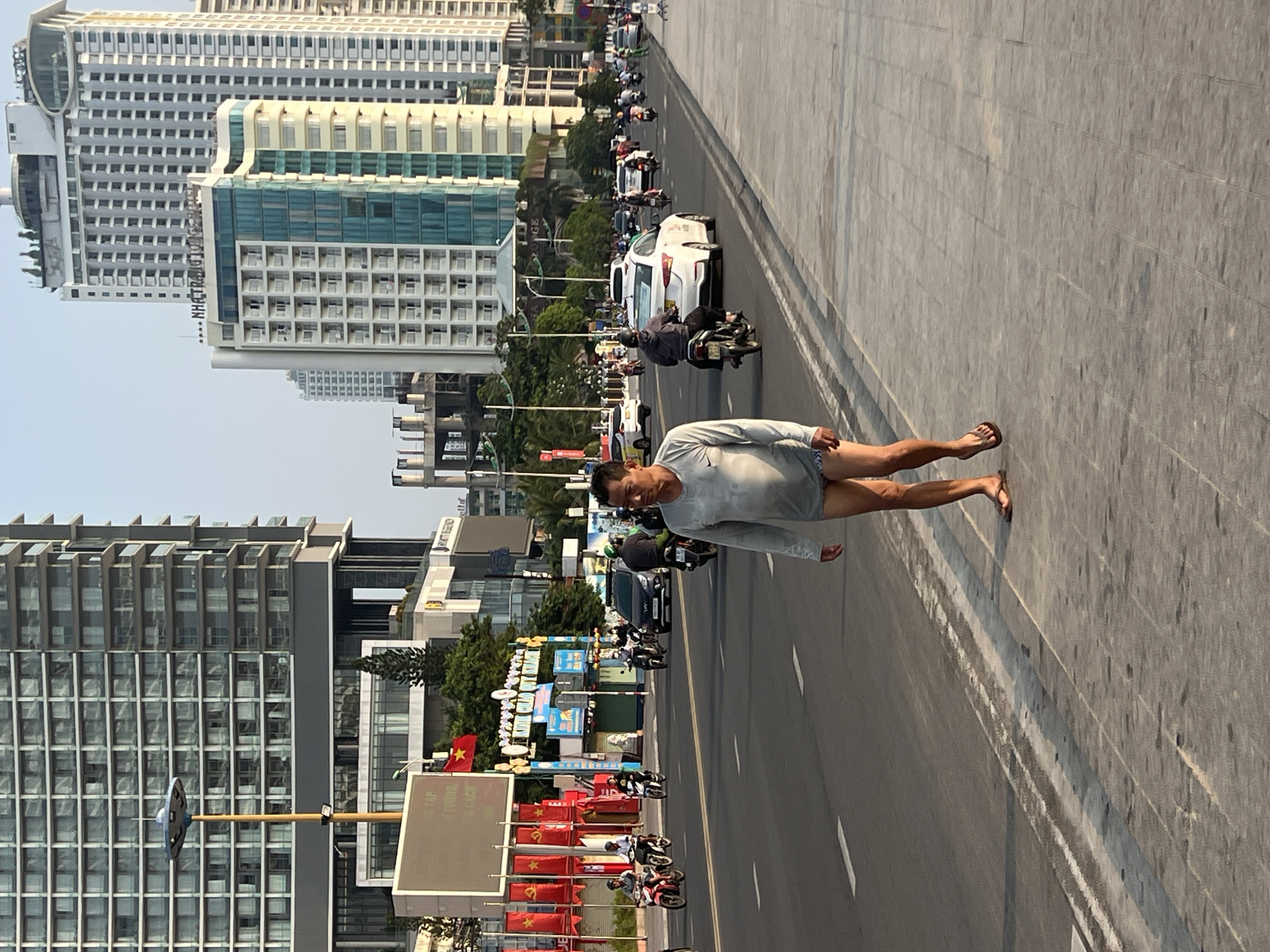 The image depicts a man walking along a concrete median on a road in Nha Trang, Vietnam. The man is wearing a light gray long-sleeved shirt and shorts, with brown sandals. He is walking on the right side of the road, facing the camera.
Behind the man, the road is filled with vehicular traffic, including motorcycles and cars, indicating a busy urban setting. Buildings, including high-rise hotels and apartments, dominate the cityscape. Vietnamese flags are visible along the median.