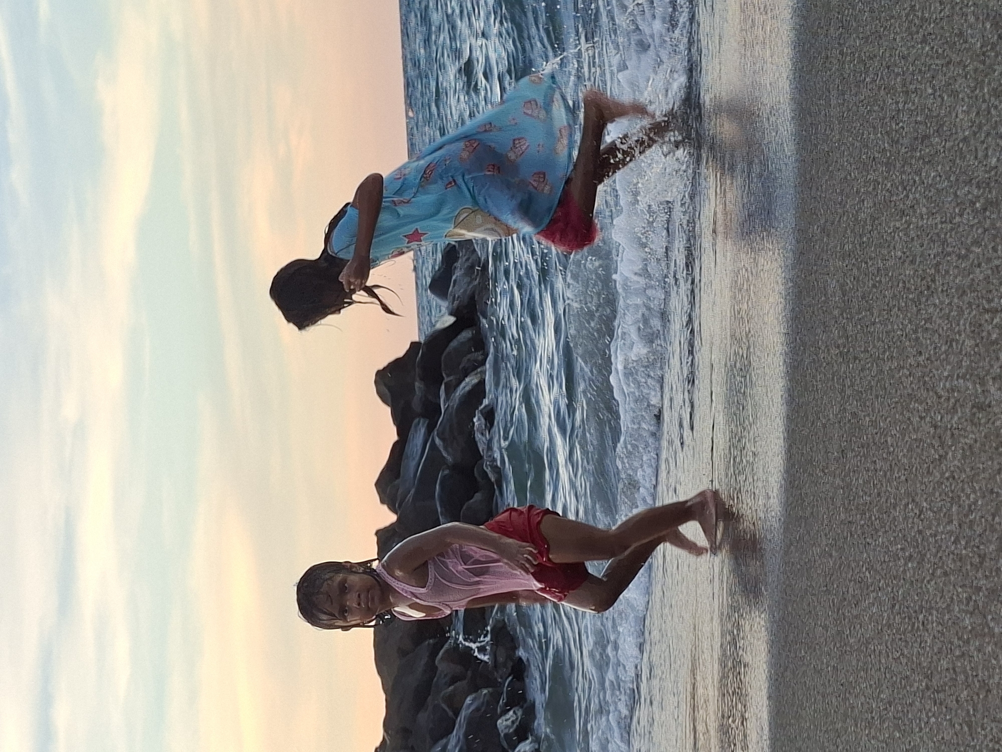 Two girls are playing on a beach in Lhokseumawe, Indonesia. One girl is wearing a blue patterned dress with pink shorts underneath and is partially submerged in the water. The other girl wears a pink tank top and red shorts, also in the water. A rocky barrier is visible in the background. The sky has an orange hue, suggesting it might be sunset. The sand of the beach extends to the right side of the image.
