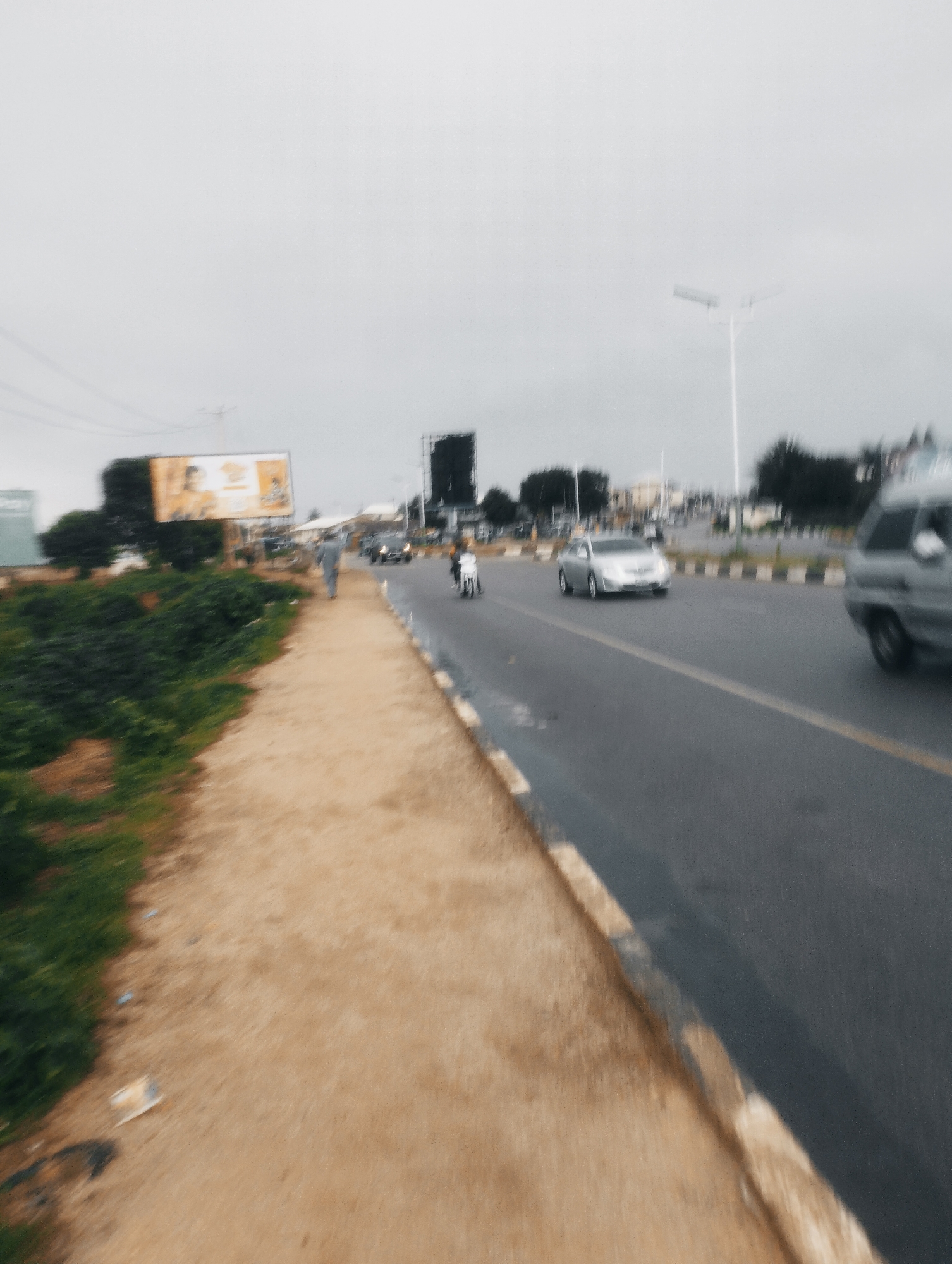 The image depicts a blurred street scene in Jos, Nigeria, characterized by a cloudy sky. In the foreground, a dusty path runs alongside a curb and a busy road. On the road, various vehicles are in motion, including a silver sedan, a motorcycle, and a light-colored van. A person is seen walking on the sidewalk, and another on a motorcycle is also visible. A billboard advertising a product is prominently displayed in the background. The overall impression is one of a typical urban environment with ongoing daily activities.