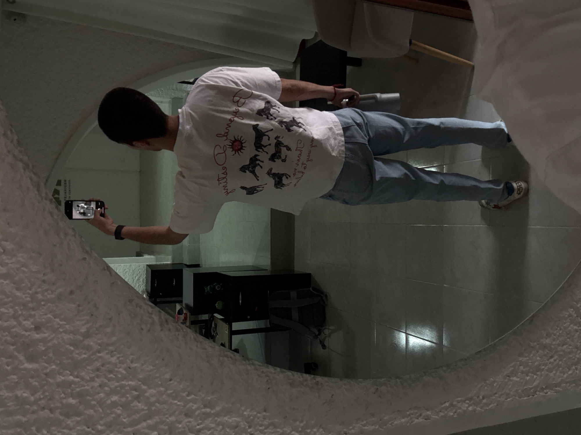 A person wearing a white t-shirt and blue jeans is taking a photo of themselves in a round mirror. The location is Puebla, Mexico. The surroundings include a tiled floor, a desk with equipment, and a white wall. The mirror's edge is textured.