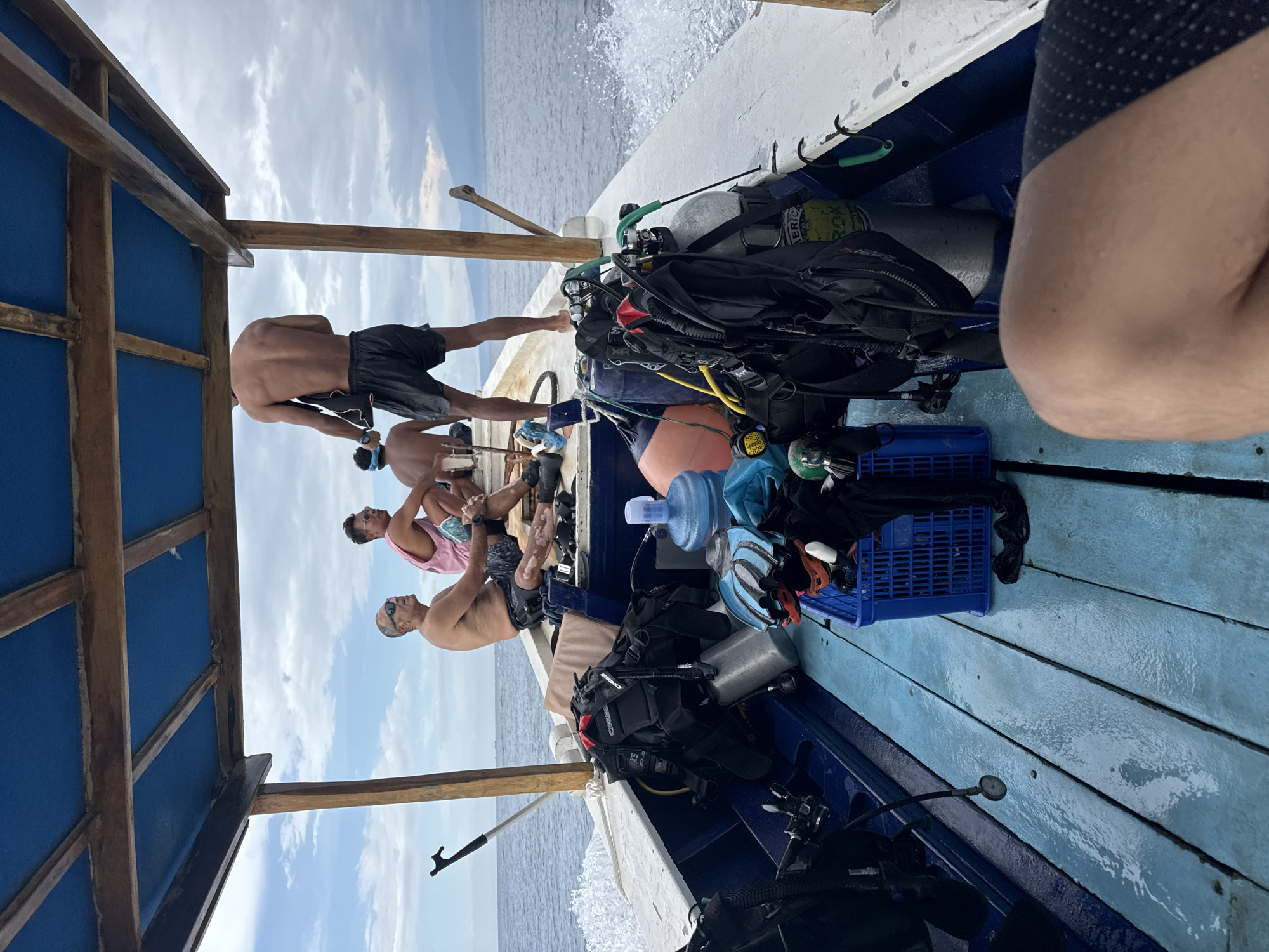 On a boat in Unknown, Indonesia, four shirtless men sit and stand near diving equipment. One man stands near the front of the boat, holding onto its side. Another man sits next to him, wearing a blue headband. Further back, a man in a pink tank top sits and looks down. Next to him sits another man with sunglasses. The boat contains numerous pieces of diving equipment, including tanks, regulators, and buoyancy compensators. A blue crate holds additional gear. The ocean stretches out behind the boat, and a cloudy sky is visible overhead.