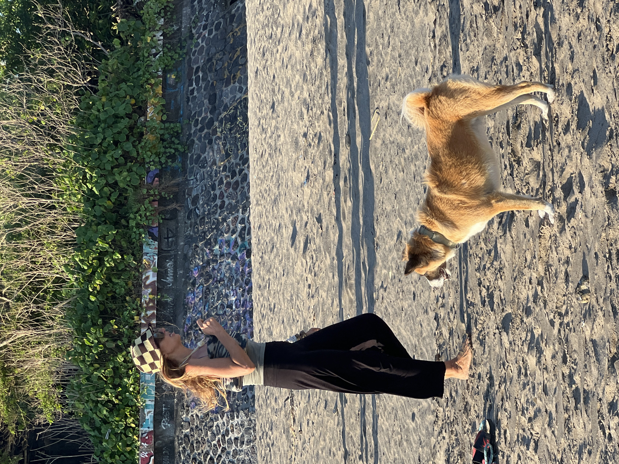 The image depicts a woman and a dog on a beach. The woman, wearing a checkered hat and dark clothing, is lying down near a wall with vegetation. A light-colored dog lies to her right, also on the beach. The scene is bathed in sunlight, casting shadows across the sand. The location is North Kuta, Indonesia.