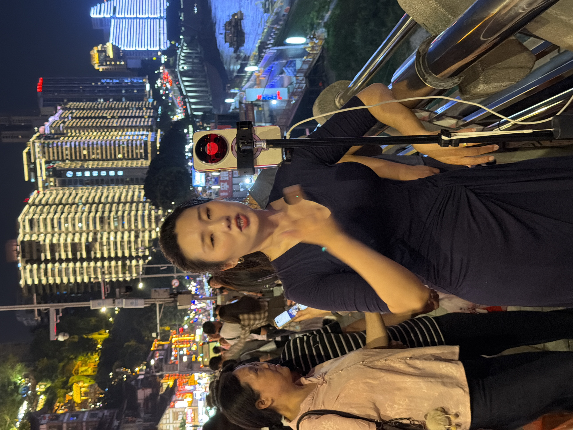 The image shows a night scene in Yuzhong District, China, with illuminated buildings in the background. A woman in a dark blue dress is in the foreground, holding a smartphone mounted on a tripod. She appears to be filming herself or taking a selfie, with her mouth slightly open as if speaking. Her expression suggests she is engaged in the activity.
Below her, another woman is visible, wearing a pink patterned top and black pants. She is looking upwards, seemingly at the woman in the blue dress or the camera. The background is a blur of city lights, indicating a bustling urban environment. The time of day is clearly night, and the lighting suggests it is a clear evening.
The overall scene captures a moment of personal recording or content creation amidst a vibrant city landscape at night. The focus is on the interaction between the two women and the technological act of filming. The presence of multiple people in the background suggests a public space, possibly a street or a promenade. No discernible text is visible in the image.