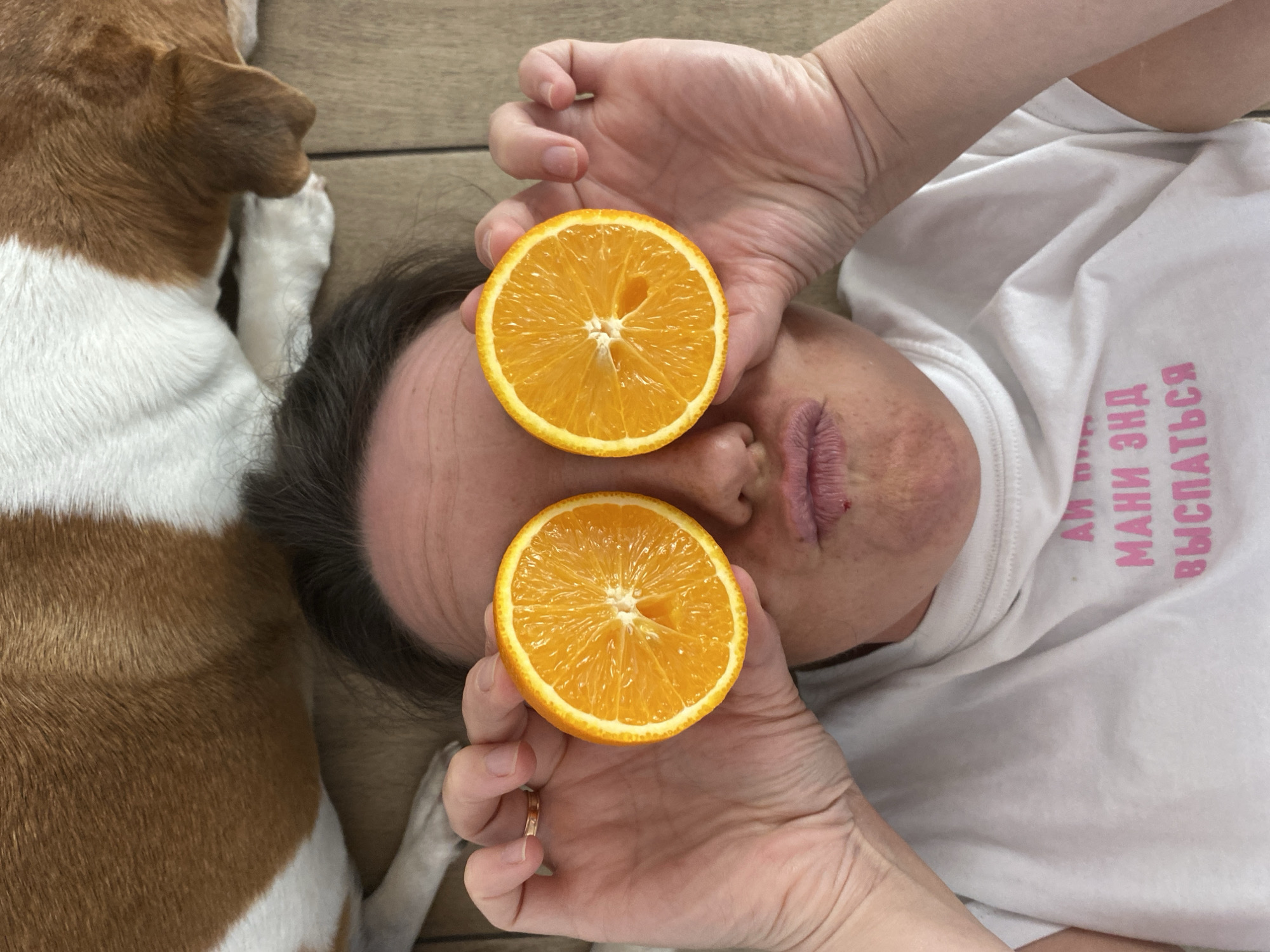 A woman lies on a wooden surface with a brown and white dog resting beside her. She holds two orange halves over her eyes. She wears a white t-shirt with pink Cyrillic text. The location is Chaikovsky, Russia.