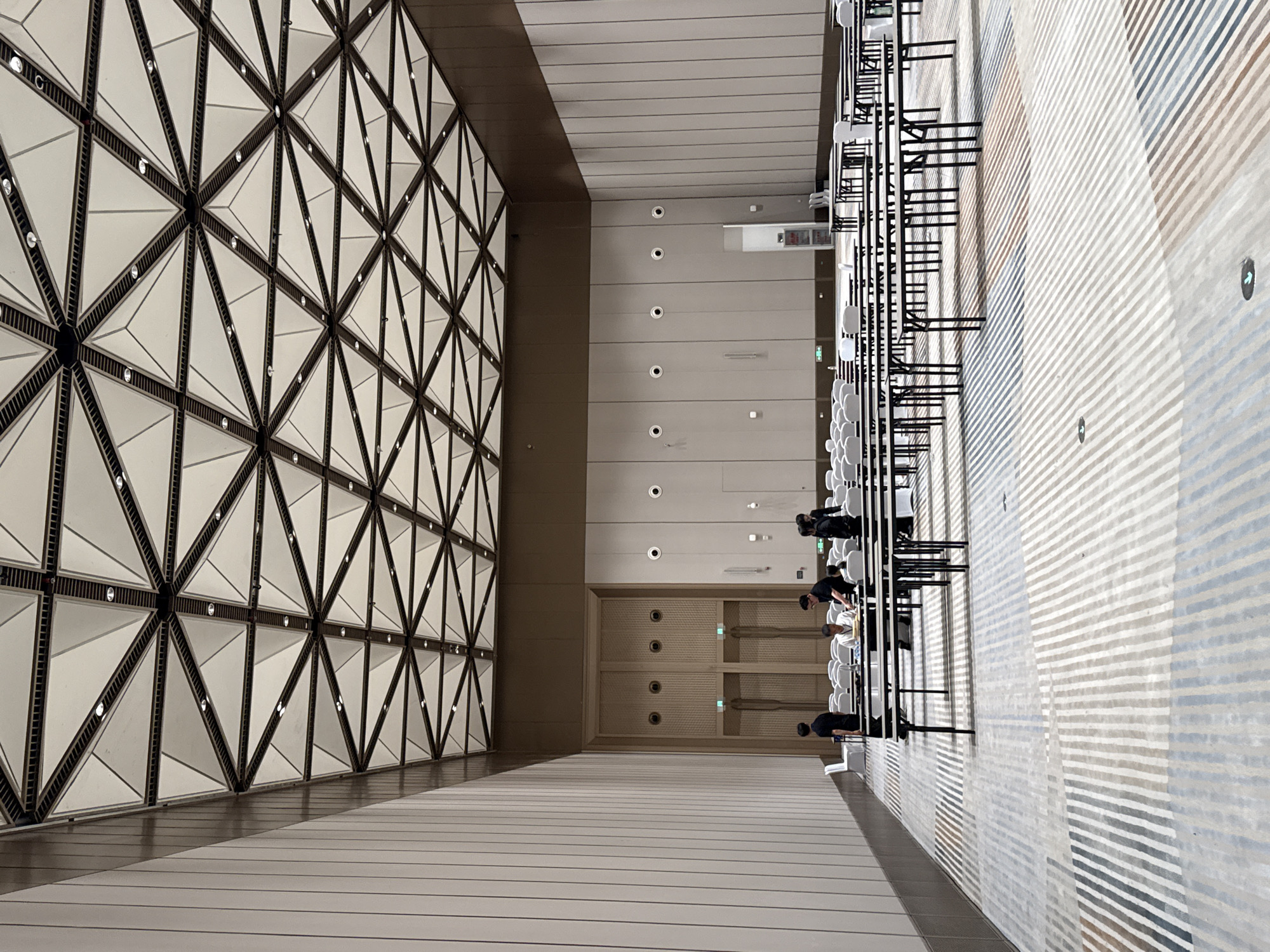 The image depicts the interior of a large, modern conference hall located in the Chongqing High-tech Zone, China. The hall features a striking geometric ceiling with triangular panels and integrated lighting. The walls are a neutral beige, with vertical paneling on the left side and circular vents or speakers on the right. The floor is covered with a patterned carpet featuring alternating stripes of white, grey, blue, and brown.
In the foreground, numerous black folding tables are arranged in rows, some with white chairs behind them. A group of four individuals are visible near a cluster of tables. Three appear to be men and one a woman, dressed in dark attire. One man is seated at a table, looking down at something, while another stands behind him, seemingly engaged in a task. The other two individuals are standing and observing.
In the background, a large set of double doors with vertical wooden slats is visible, flanked by what appear to be additional doors or entrances. Rows of empty white chairs are set up behind the tables, suggesting preparations for an event. The overall impression is one of a spacious, well-lit venue ready to host a gathering.
