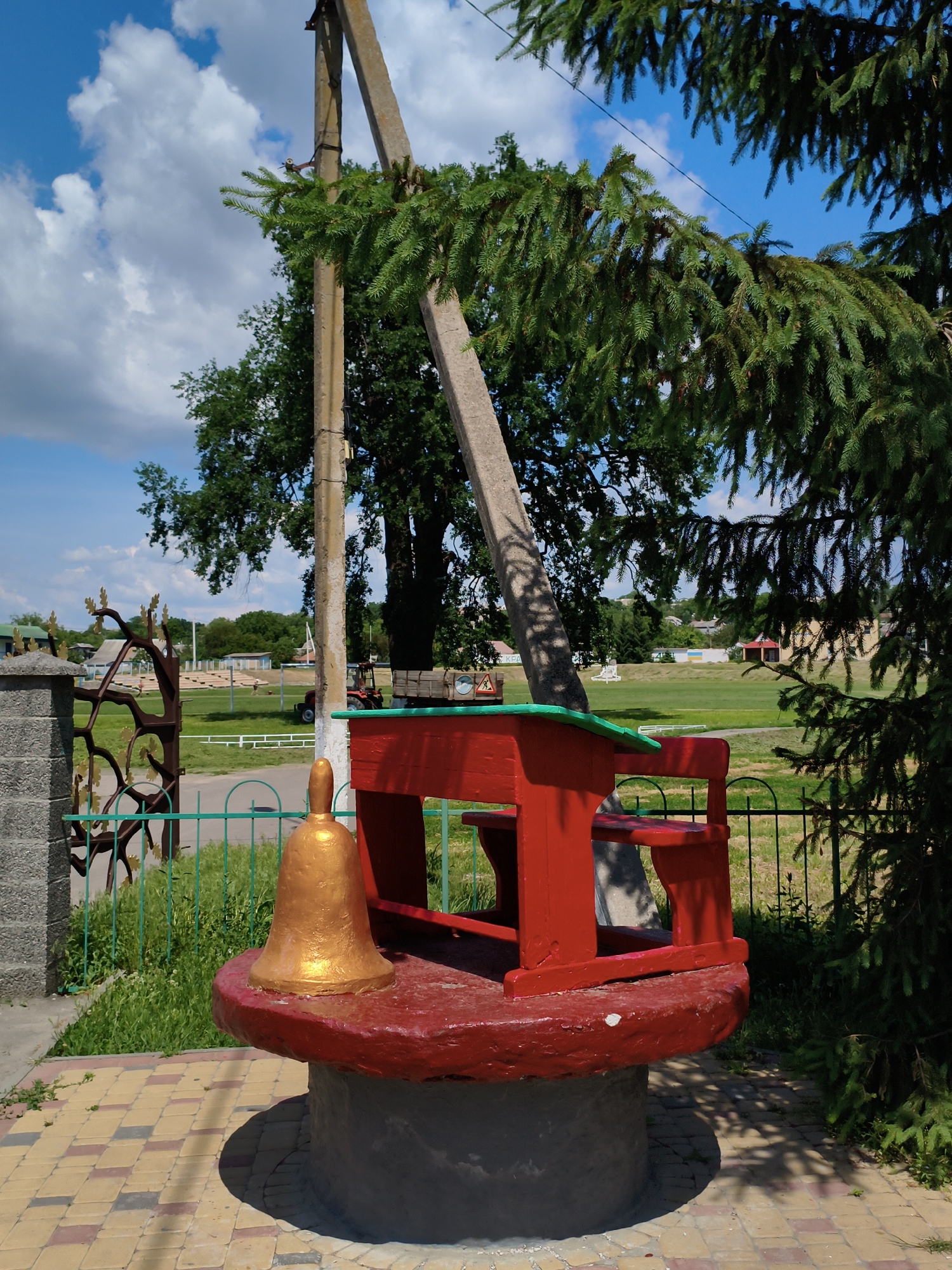 The image captures an outdoor scene in Velyka Bahachka, Ukraine, featuring a decorative red school desk and bell structure situated on a circular, red-painted base atop a concrete cylindrical foundation. The structure is adorned with a green accent along the desk's edge and a golden bell with a clapper is placed prominently on the red base. Behind the desk, a large concrete pole supports a leaning, angled concrete support beam, partially obscured by the lush green branches of an adjacent evergreen tree. A metal fence with vertical green bars encloses the area in front of the structure, and beyond the fence lies a grassy field. A decorative metal gate with a branch motif is visible to the left, next to a grey stone pillar. In the background, trees and buildings can be seen, and a red tractor is parked in the field. The sky above is blue with scattered white clouds. The ground in the foreground is paved with a pattern of yellow and reddish-brown interlocking bricks.