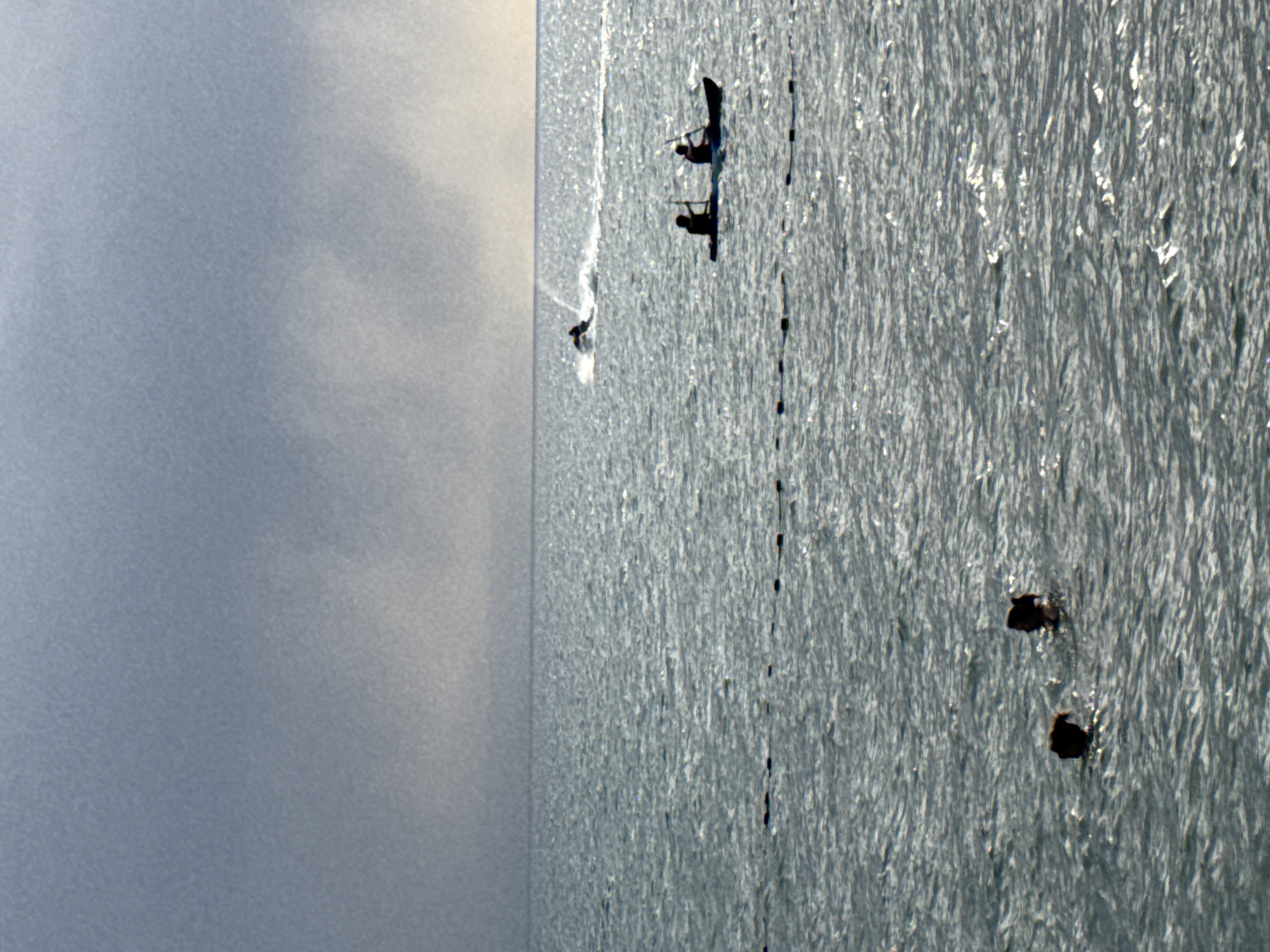 In Sary-Oy, Kyrgyzstan, on a body of water under a grey, overcast sky, two individuals are visible swimming in the foreground. Further out in the water, two people are seated in a kayak, paddling. In the background, a jet ski is being operated by a single person, leaving a wake behind it. The water's surface is choppy and reflects sunlight. A line of buoys or markers is visible in the mid-ground, stretching horizontally across the frame.