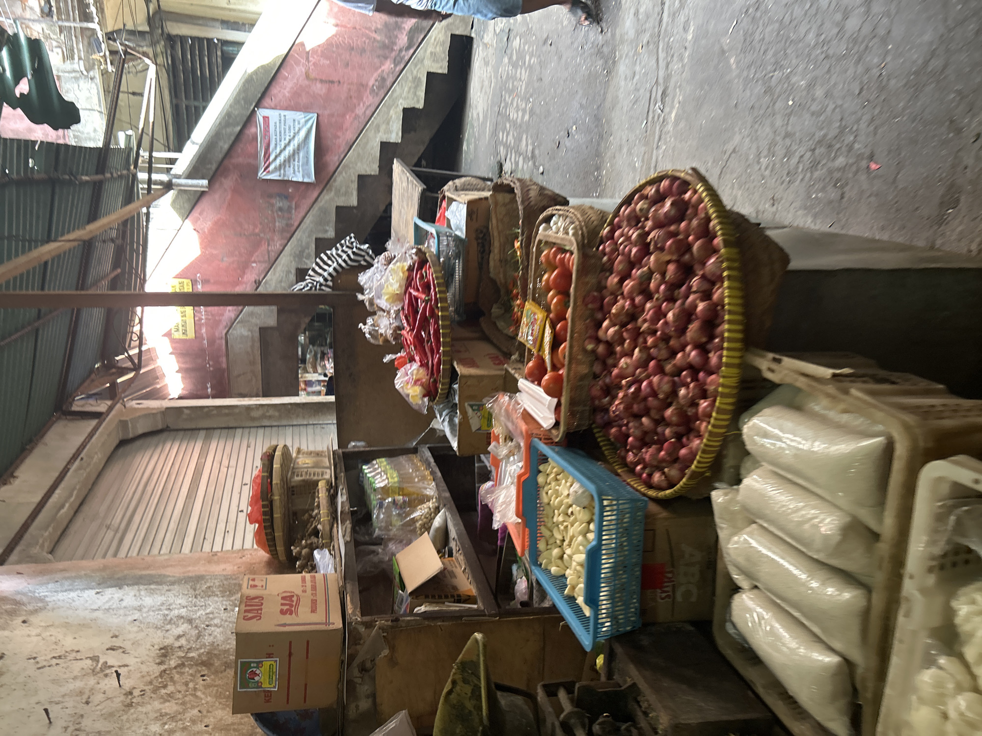 This image depicts an outdoor market stall in Denpasar, Indonesia. The stall is filled with fresh produce, including a large basket of red onions in the foreground, a basket of garlic cloves, and a container of tomatoes. Red chilies are also visible in a woven basket. Bags of rice are neatly arranged in plastic containers. Behind the produce, shelves display various goods, including boxes, bottles of liquid, and other food items. The market appears to be set up under a covered roof, with a concrete floor. In the background, a red staircase and a rolling metal door suggest the market is situated within a more complex structure, possibly a building or a covered market area. The overall impression is of a bustling marketplace, typical of local commerce in Southeast Asia.