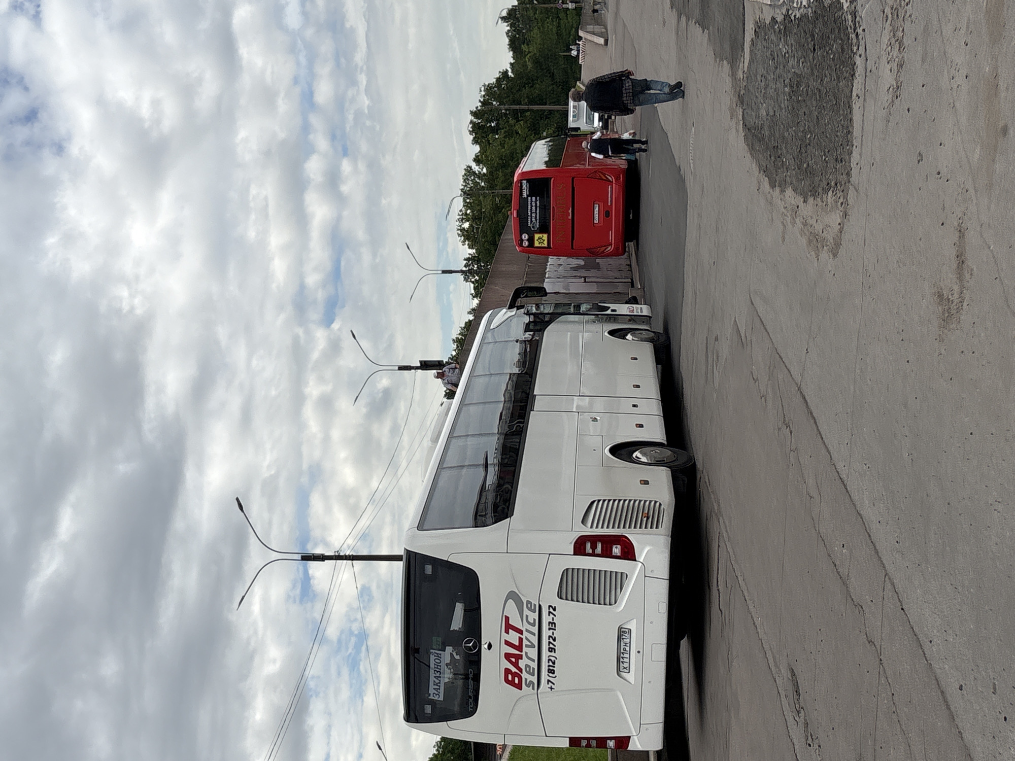 The image depicts a street scene in Saint Petersburg, Russia, featuring two buses. The foreground is dominated by the rear of a white coach labeled "BALT SERVICE" with a Russian license plate that reads "X111PH 178". The bus also displays "ЗАКАЗНОЙ" which translates to "booked" or "chartered." To the right, a red bus is parked, also with Russian signage. Several people are visible in the scene: two men are walking away from the camera towards the red bus, and another person, likely a man, is standing on top of the white bus. In the background, trees and a cloudy sky are visible, along with streetlights and what appears to be a retaining wall. The ground is paved with asphalt and shows signs of wear and tear.