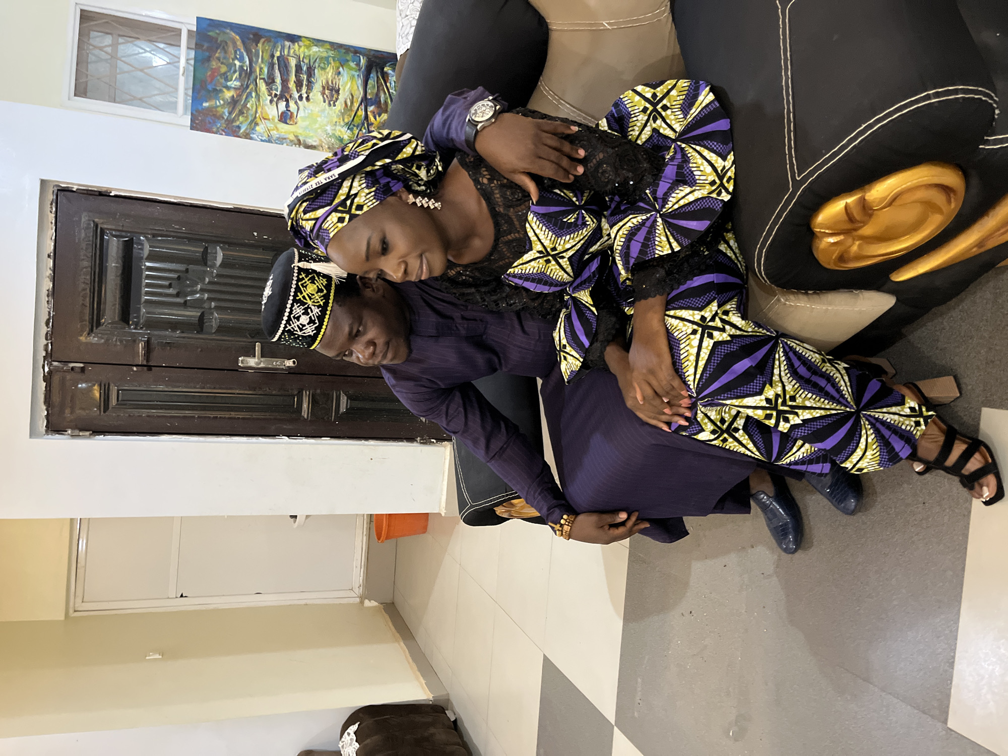 A couple is sitting closely together on a couch. The man, wearing a black hat with gold embroidery and a dark blue suit, is seated with his arm around the woman. The woman, wearing a purple and gold patterned outfit with a matching headwrap, is leaning against him. They appear to be indoors, with a tiled floor and a painting visible on the wall. The setting is Jos, Nigeria.