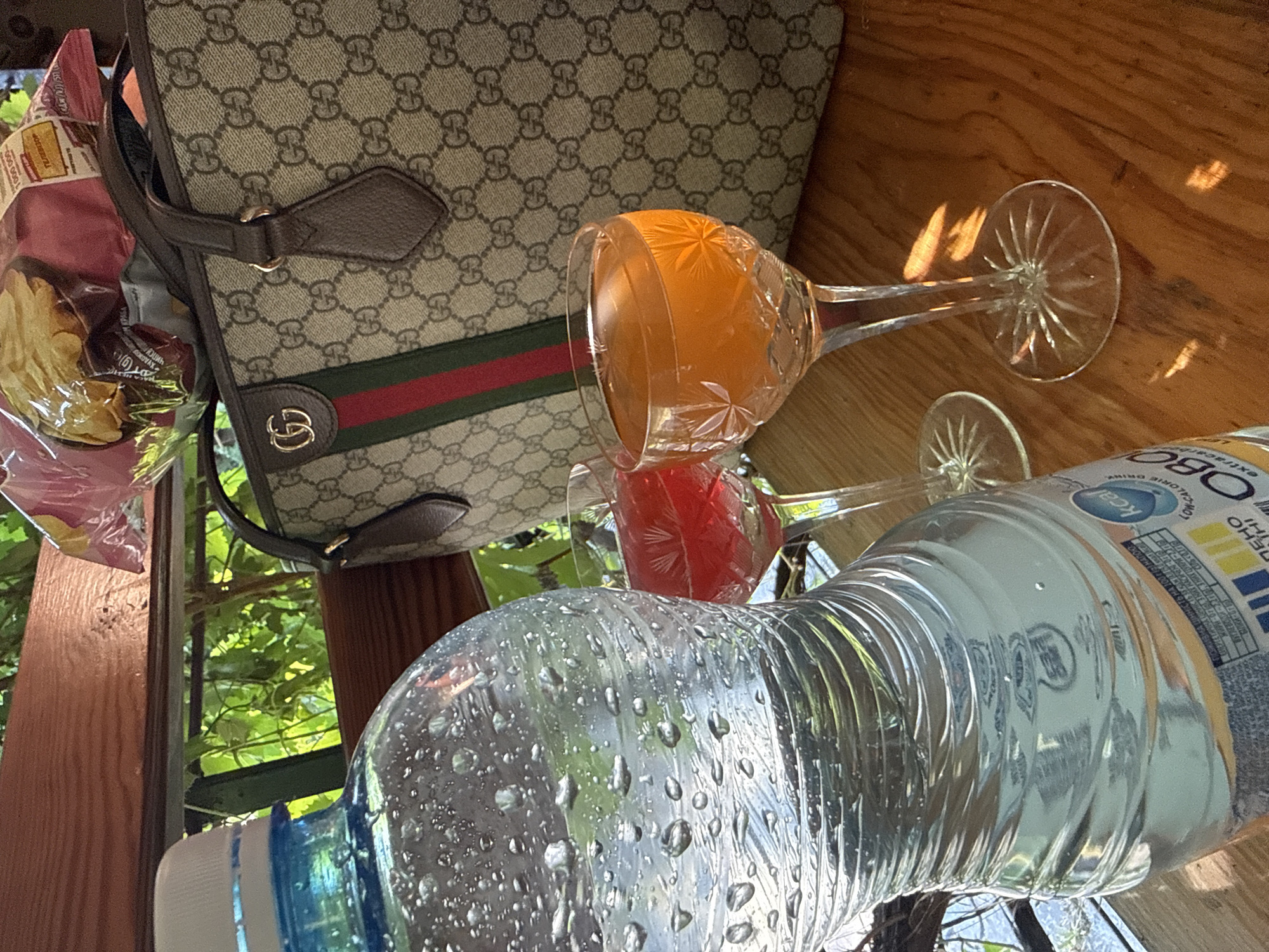 The image depicts an outdoor setting, likely a cafe or restaurant, with a wooden table in the foreground. On the table, there is a large plastic bottle of water with condensation on its surface. Next to the bottle are two stemmed glasses, one filled with a red beverage and the other with an orange beverage. In the background, a designer handbag with the Gucci logo is visible, along with a bag of snacks. Green foliage and a wooden structure suggest an outdoor, possibly shaded, environment. The scene is well-lit, with reflections visible on the table and glassware. The image does not provide specific clues to pinpoint its exact location within the city of Obukhivka, Ukraine, beyond the general context of an outdoor seating area.