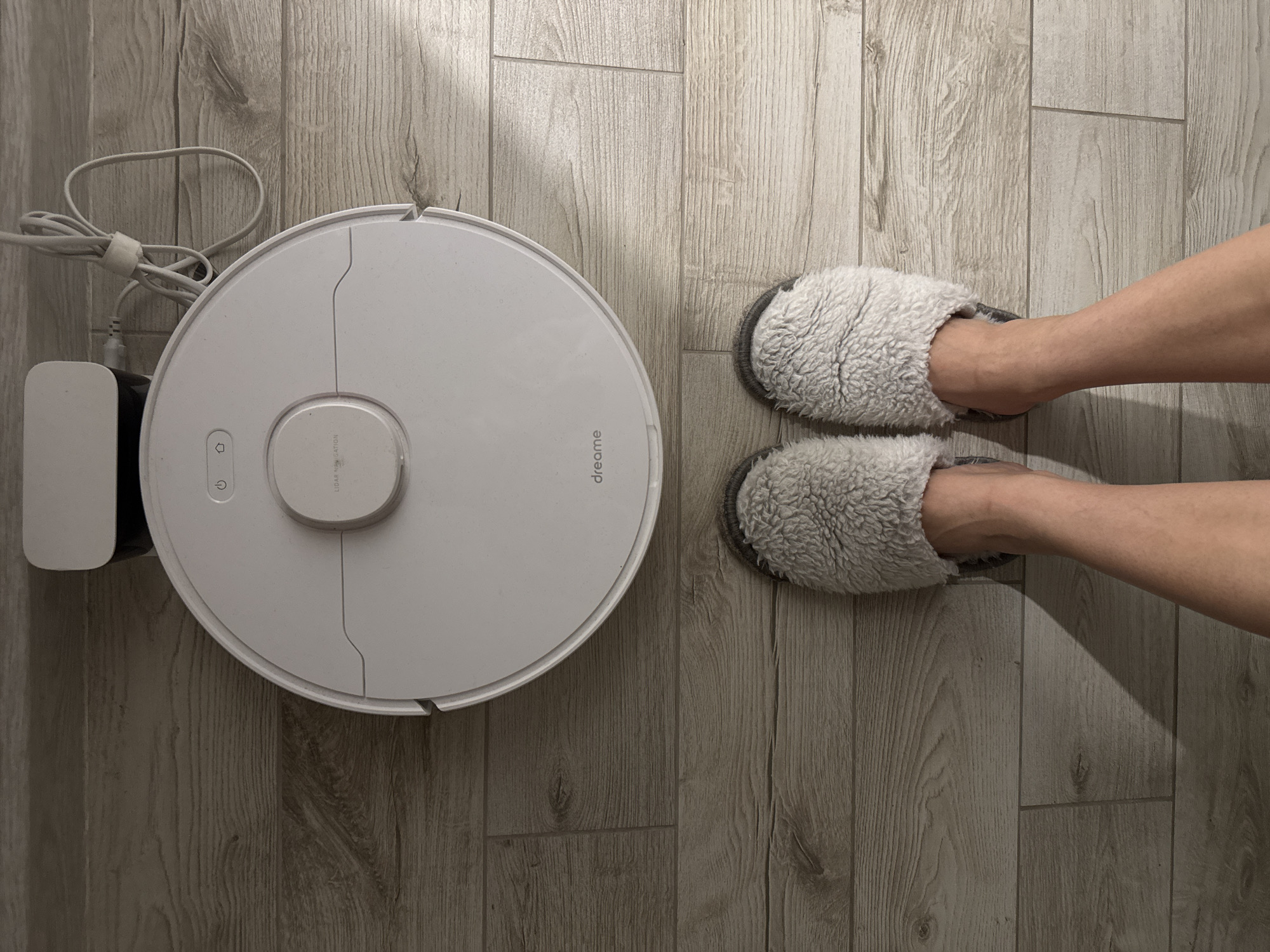 A top-down shot depicts a domestic scene. A white, circular robot vacuum cleaner (possibly a Dreame model, as the brand is visible on its surface) sits to the left with its charging station and cord. To the right are two feet wearing light gray, furry slippers. All elements are placed on a light gray, wood-grain-patterned floor. The location and city cannot be determined from this image.