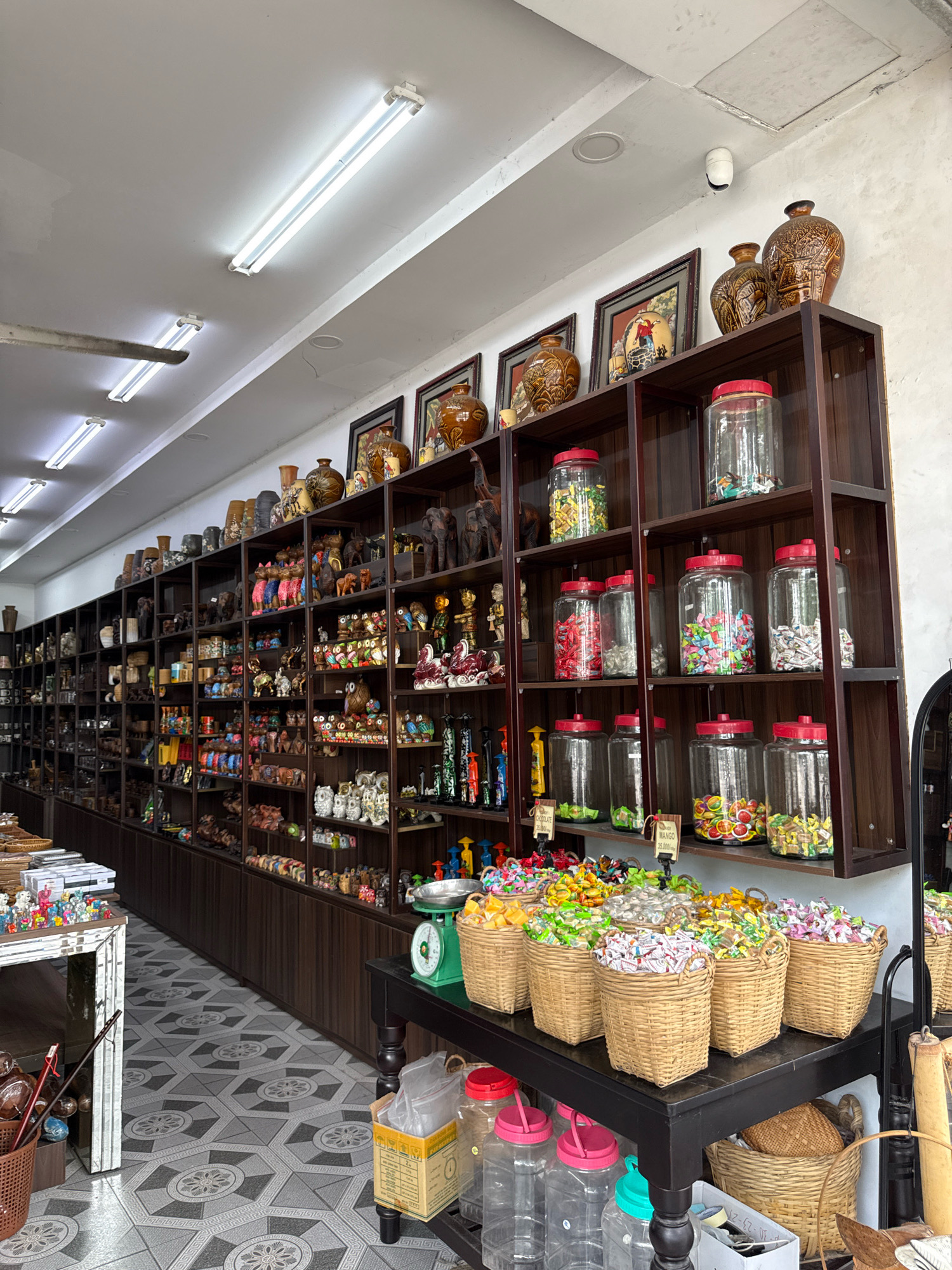 This is a wide shot of an interior space, possibly a shop in Nha Trang, Vietnam. The scene is dominated by dark wooden shelves filled with a variety of items. On the upper shelves, there are numerous ceramic vases and framed pictures. Below these, the shelves are packed with rows of small decorative objects, including what appear to be wooden figurines of animals like elephants and owls, as well as other trinkets.
In the foreground, a dark table holds an assortment of woven baskets filled with individually wrapped candies in various colors, suggesting a confectionery or gift shop. To the left of this table, a white table with decorative elements displays more small figurines. A green vintage scale is also visible on the dark table. Large clear glass jars filled with candies are situated on the shelves above the baskets. The floor is covered with a geometric tile pattern in shades of grey. Overhead, fluorescent lights illuminate the space. There are no people visible in the image.