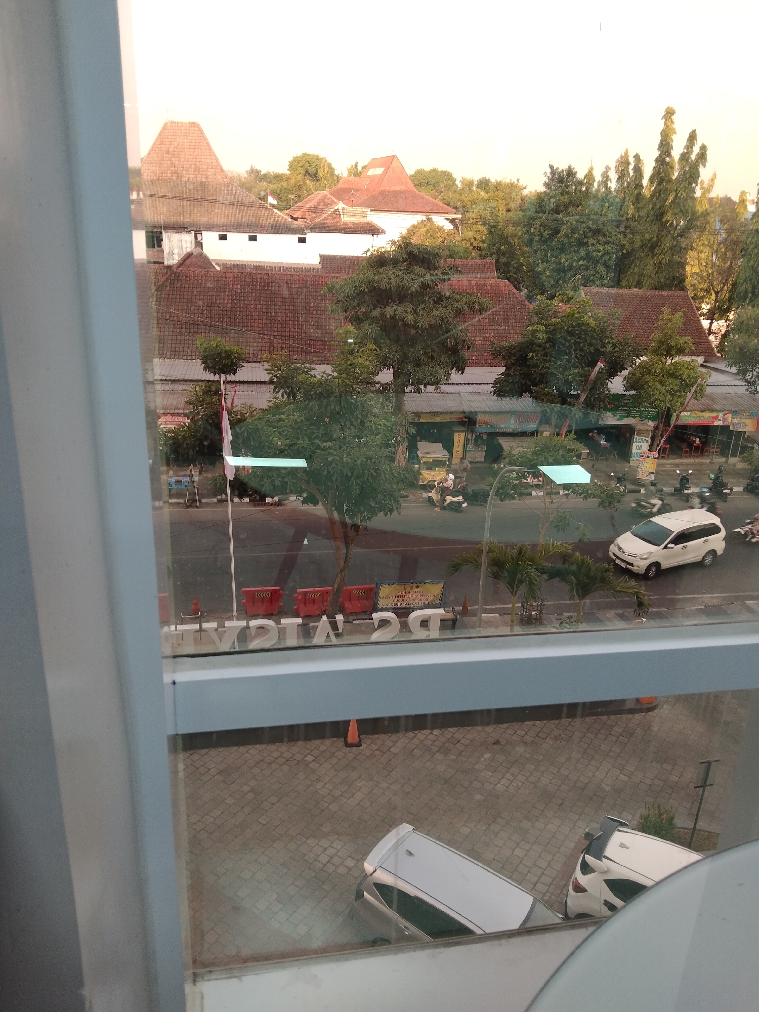 The image shows an aerial view of a street in Bojonegoro, Indonesia, as seen through a window. In the foreground, part of a building's interior is visible, including a window frame and what appears to be a white wall. The window looks out onto a street with traffic, including cars and motorcycles. Several buildings with red tile roofs are visible on the side of the street, some with signage that is partially obscured. Trees and foliage are also present throughout the scene. The sky is a pale yellow, suggesting it is either sunrise or sunset. In the foreground on the street level, there are some orange traffic cones and red barriers. Further back, several parked cars, including white sedans and SUVs, are visible in a paved area. The overall scene depicts a typical urban environment in Indonesia.
