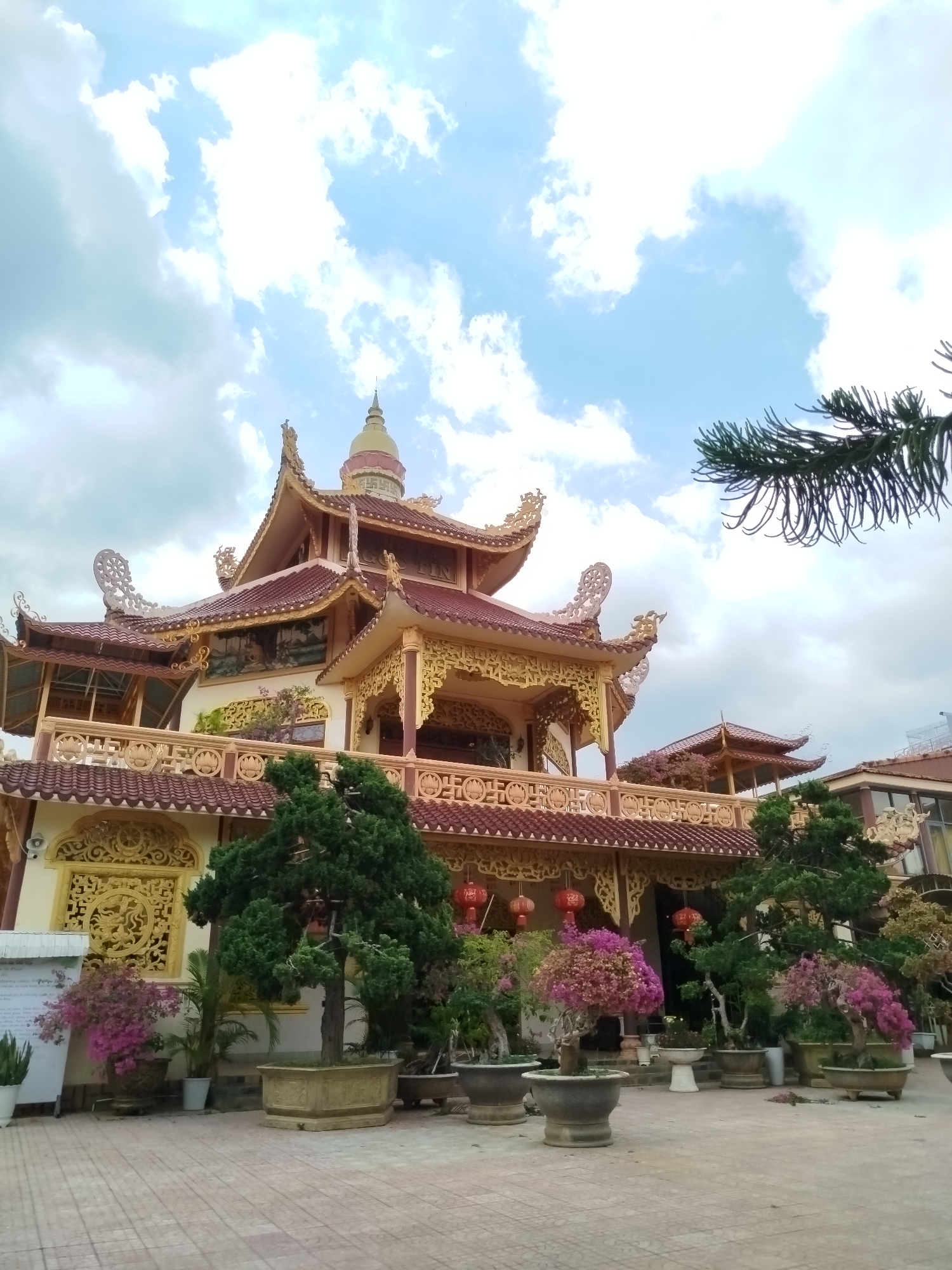 The image shows a Buddhist temple in the city of Lâm Viên - Đà Lạt, Vietnam. The temple is a multi-tiered structure with ornate gold and red details. In the foreground, there are several potted plants and trees, including bonsai trees with pink flowers. The ground is paved with square tiles. The sky is cloudy with patches of blue. A branch of a pine tree is visible in the upper right corner. Red lanterns hang from the temple's eaves.