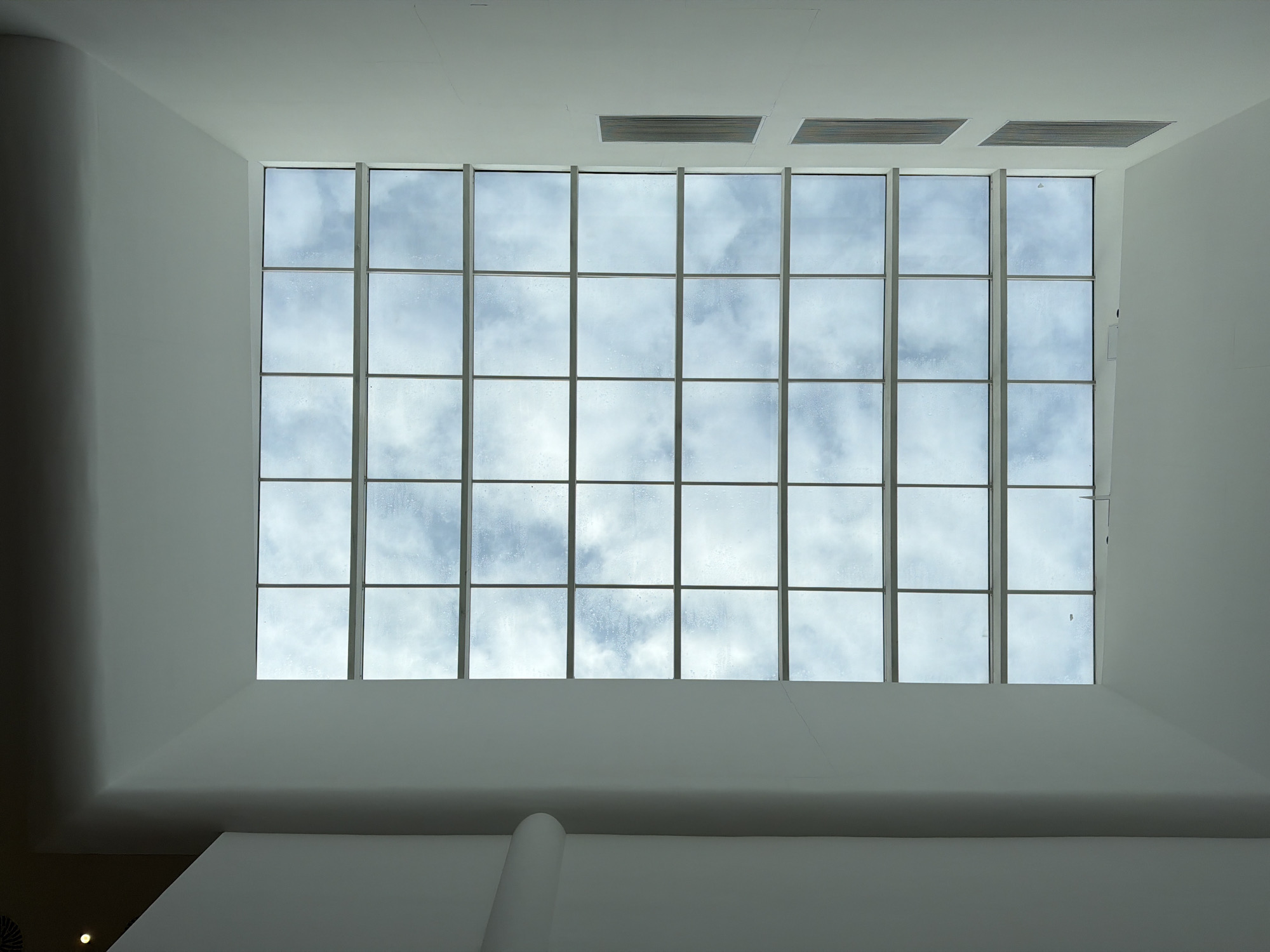 A low-angle interior shot depicts a large, rectangular window divided into 24 panes, revealing a cloudy sky. The window is set within a white wall structure. Ventilation grilles are visible above the window. The scene is devoid of people. The location is not discernible as Moscow, Russia, from the given image.