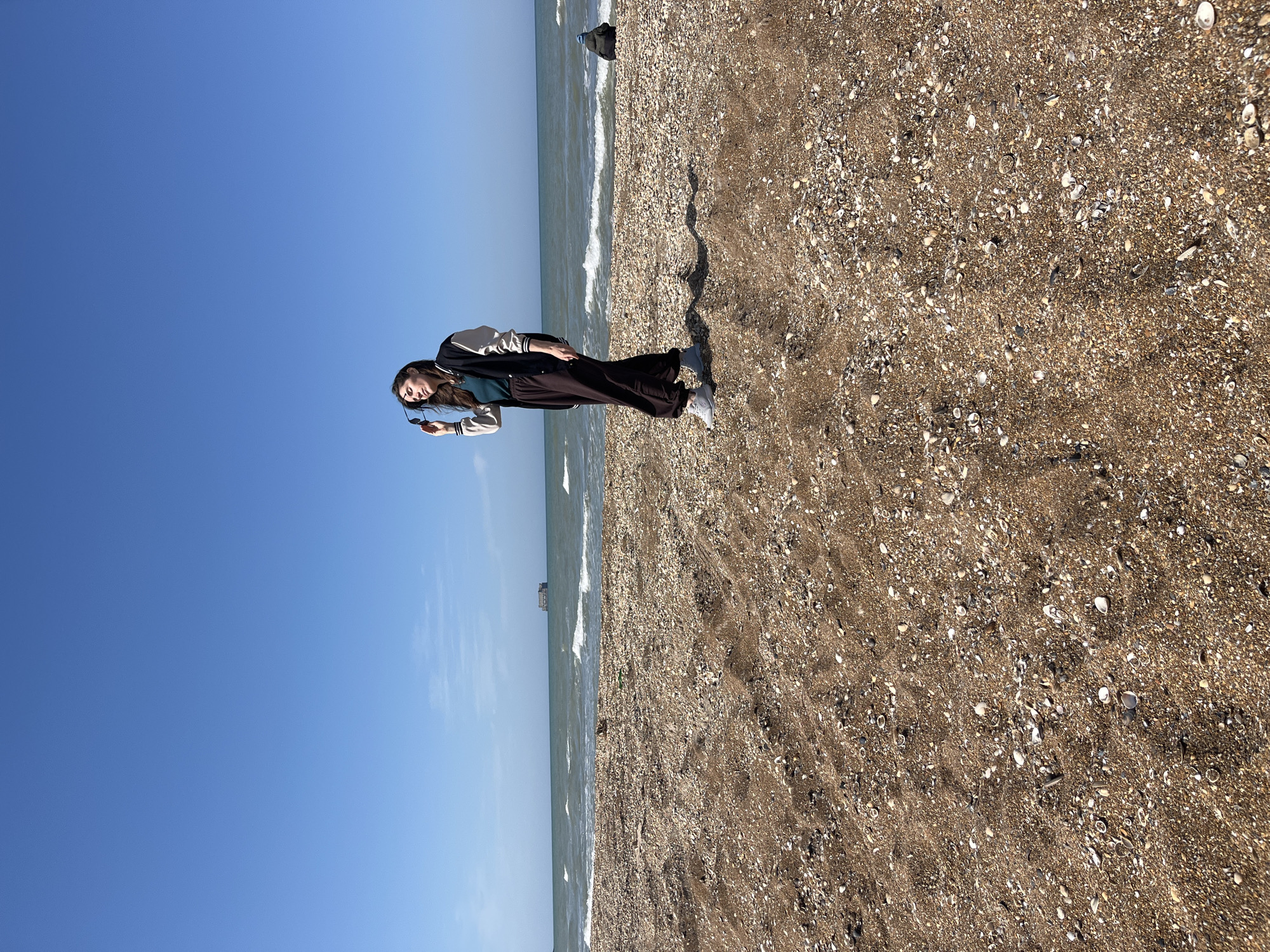 A young woman stands on a beach in Kaspiysk, Russia. The beach is composed of brown sand and shells. The sea is visible in the background, meeting a clear blue sky. The woman is wearing a jacket, dark pants, and sneakers. She is holding sunglasses in her left hand. A pier-like structure can be seen in the sea's distance. The scene is lit by natural sunlight.
