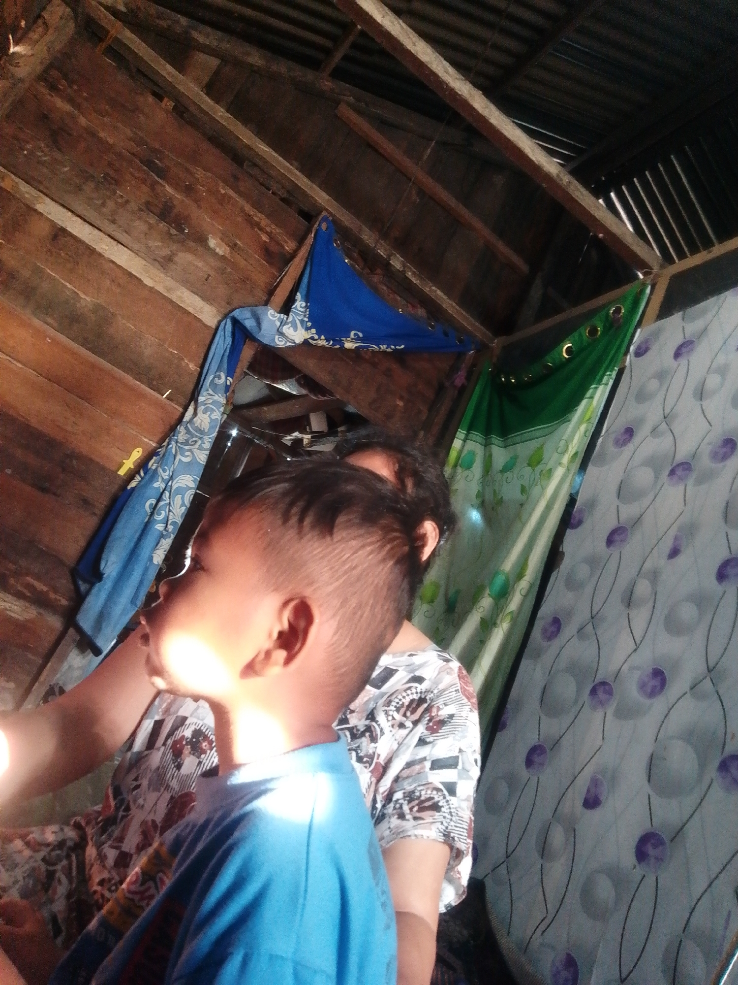 The image captures a young boy in the foreground, wearing a blue t-shirt, looking upwards with a sunlit face. Behind him, an adult's shoulder and head are visible, with the adult wearing a patterned shirt. The setting appears to be an indoor space with a rustic, unfinished wooden ceiling and walls. Hanging from the ceiling are pieces of fabric: a blue one with a white pattern on the left and a green one with grommets and a floral pattern on the right, partially obscuring a wall covered with a patterned fabric that has circles and lines. The overall impression is of a simple, possibly rural, dwelling. The provided context indicates the location as Labuhan Bilik, Indonesia, suggesting this scene is within a household in that area.