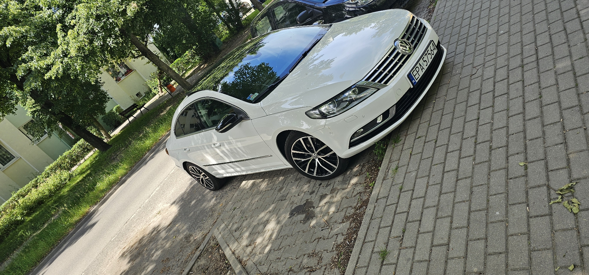 The image shows a white Volkswagen CC parked on a brick-paved sidewalk next to a road. The car has a black roof and tinted windows. The license plate is visible and reads "EPA 57564". The car is parked alongside a green grassy area with trees and a building in the background. The building appears to be a residential block with windows and some greenery on its facade. The trees are lush and full of leaves, suggesting it is daytime and likely a warm season. Sunlight casts shadows on the sidewalk and road, indicating a sunny day. The scene is set in Pabianice, Poland. There are no people visible in the image. The overall impression is a quiet, everyday street scene.