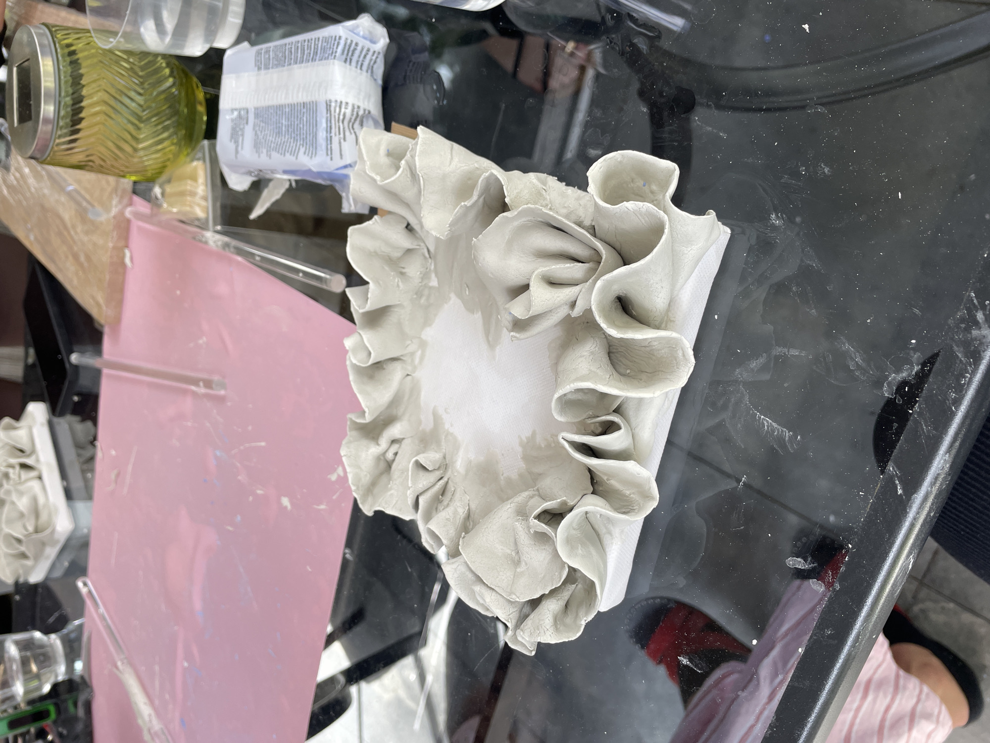 The image depicts a close-up of a square-shaped, light gray ceramic piece with a ruffled edge, resting on a glossy black surface. The ceramic piece is made of a pliable material, possibly clay, with visible folds and undulations along its perimeter, creating a decorative, organic form. The interior of the piece is white and appears to be a different material, possibly paper or fabric. The background shows a blurred environment with various objects on a table, including a pink mat, clear acrylic rods, a green glass jar, and a package of white material. The overall lighting suggests an indoor setting, likely a workspace.