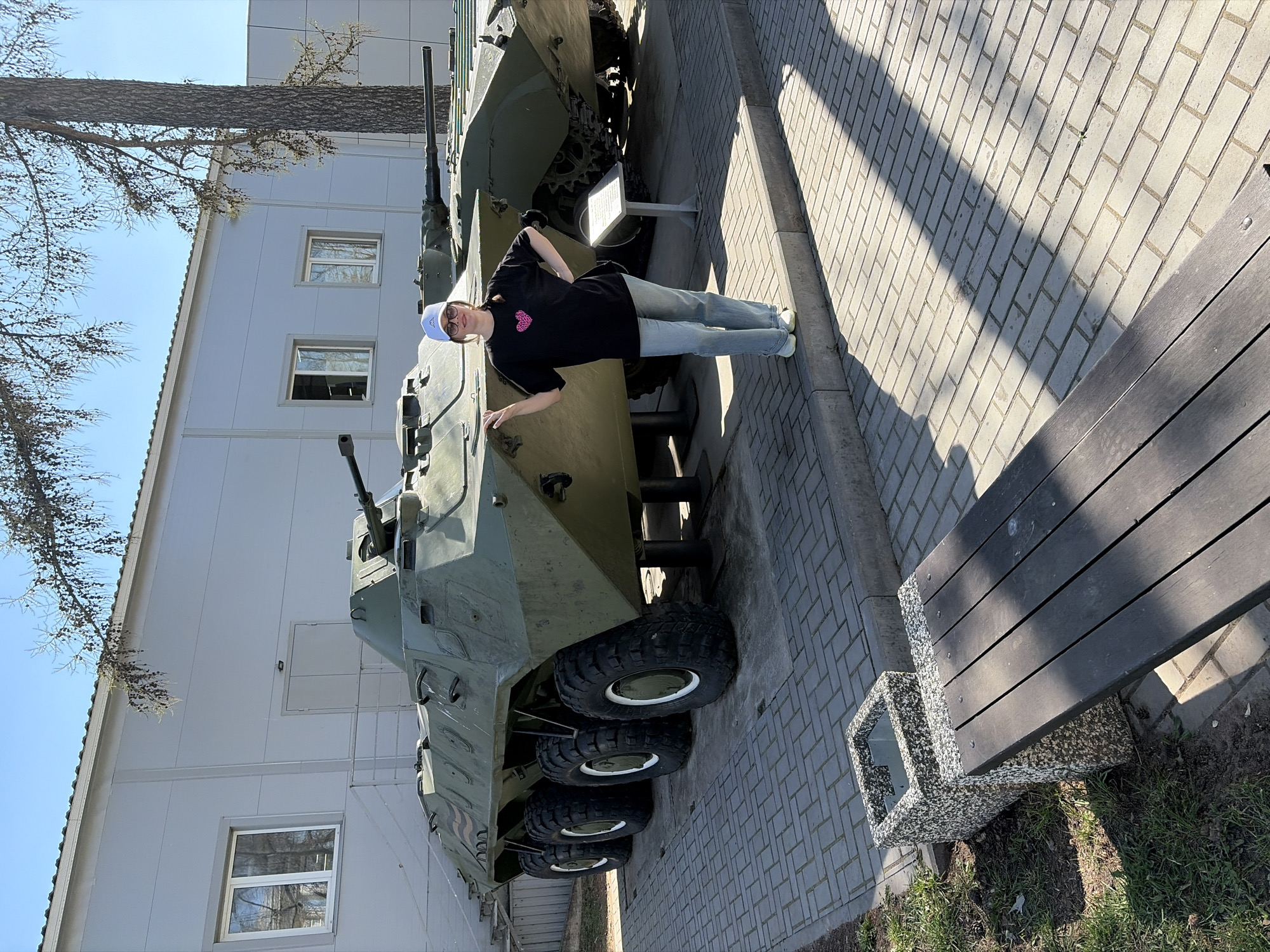 A young woman stands next to a BTR-80 armored personnel carrier in Votkinsk, Russia. The APC is olive green and has multiple wheels visible along its side. A small white sign stands next to the vehicle. The woman is wearing a black t-shirt with a pink heart, blue jeans, and a white baseball cap. She is standing on a brick-paved walkway. To the right of the walkway is a bench and a small patch of grass. In the background is a light gray building with windows and a tree with bare branches extending towards the sky. The sky is blue and clear.