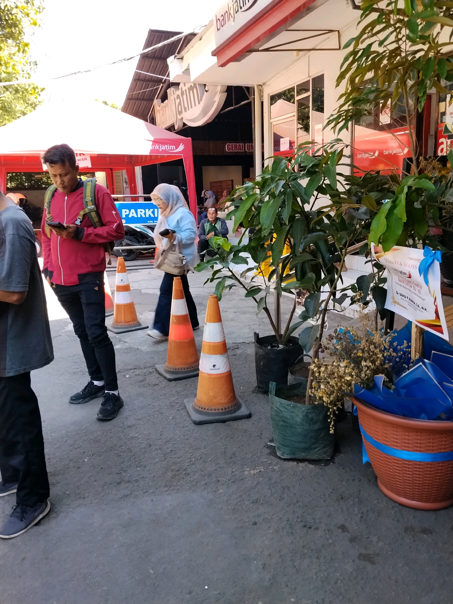 The image depicts an outdoor scene in front of a Bank Jatim branch in Bondowoso, Indonesia. A man wearing a red jacket and black pants is looking at his phone, with a backpack on. Next to him, a woman in a light blue hijab and grey pants is walking and holding a purse. Several orange traffic cones are placed on the ground, indicating a queue or a restricted area. In the background, there's a tent with "Bank Jatim" branding and a building with windows. Plants in pots are also visible, along with a blue ribbon tied to a certificate. The ground is paved asphalt.