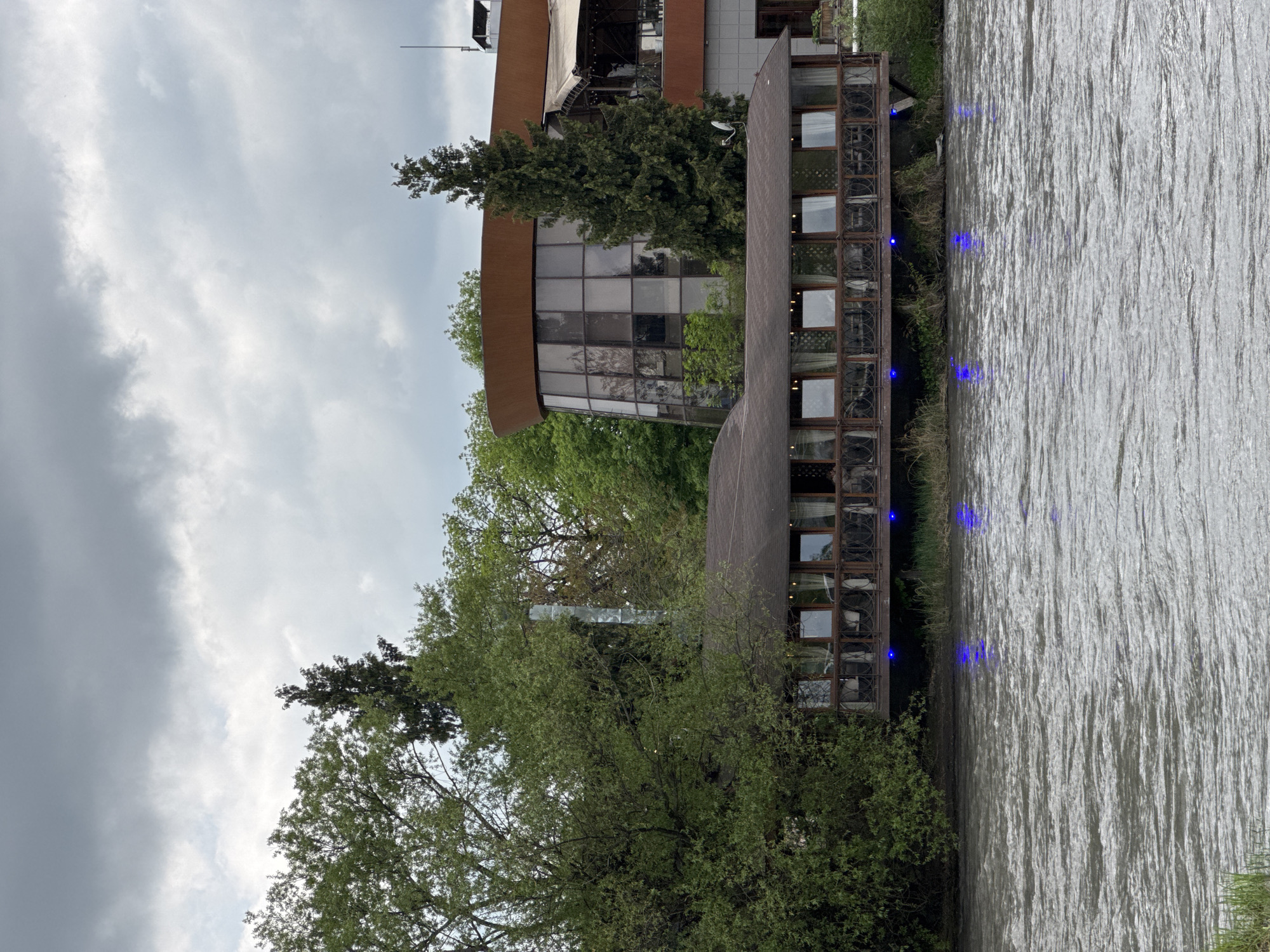 This image shows a multi-level building complex on the edge of a body of water in Zamankul, Russia. The architecture incorporates large windows and a cylindrical design element, partially obscured by trees. The sky is overcast with gray clouds. Ripples are visible on the surface of the water, and blue lights illuminate the side of the building closest to the water.