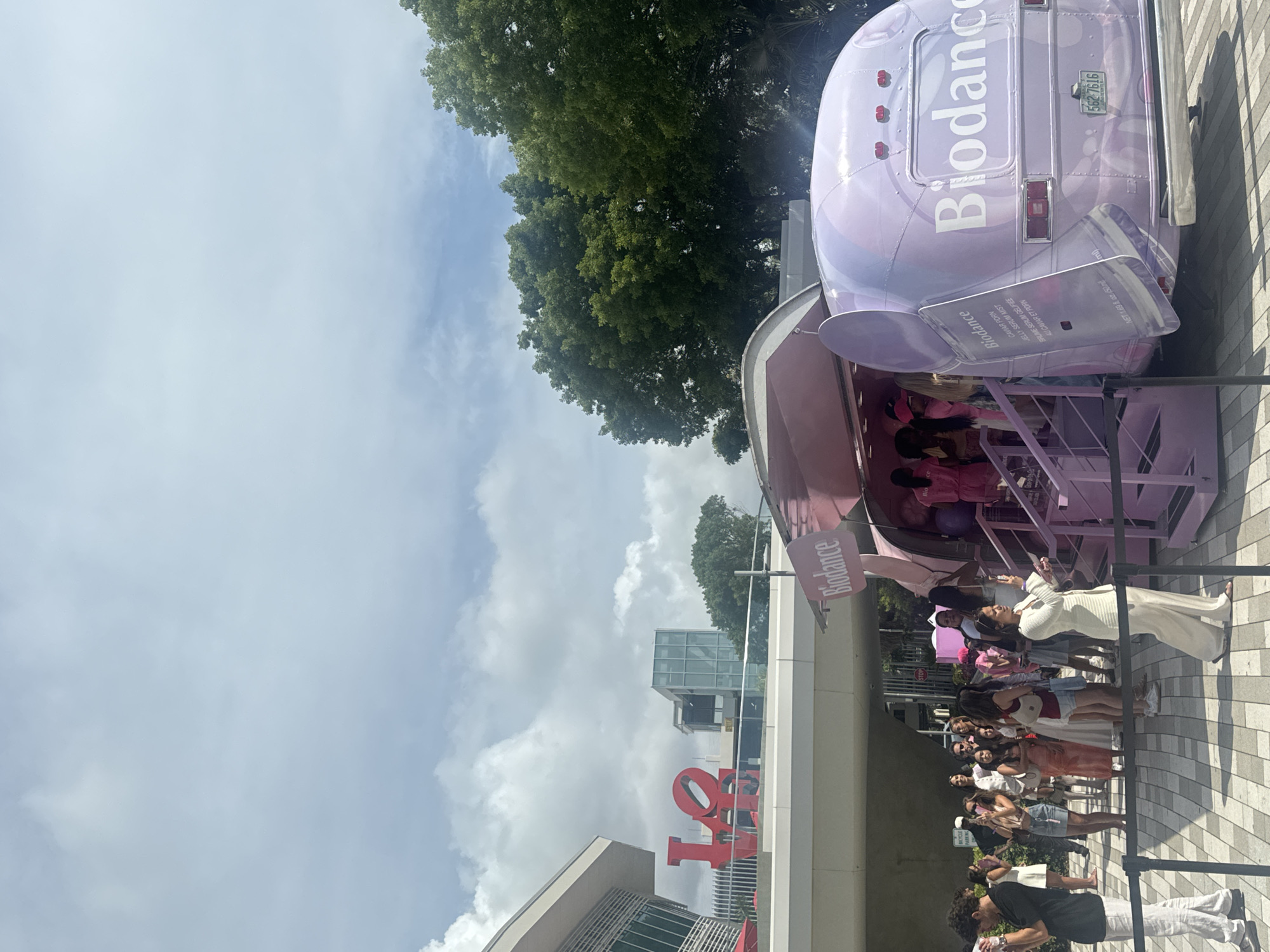 An outdoor scene depicts a crowd of people gathered around a purple mobile unit that resembles a bus. The unit is branded with the name "Biodance" in large white letters. The bus has its side opened, revealing steps leading up to an interior, where several individuals are visible. The exterior of the bus also features prominent branding and what appears to be product packaging graphics. License plate information on the back of the bus reads "56Z 7616 FLORIDA". In the background, a large red sculpture spelling out "LOVE" is visible, situated on a walkway with a modern architectural building behind it. Lush green trees and a cloudy sky complete the environment. The people in the foreground and around the bus are dressed in casual attire, with some holding phones, suggesting they are attending an event or promotional activity. The setting appears to be a public space in Aventura, United States of America, likely an outdoor plaza or walkway adjacent to a commercial or entertainment venue.