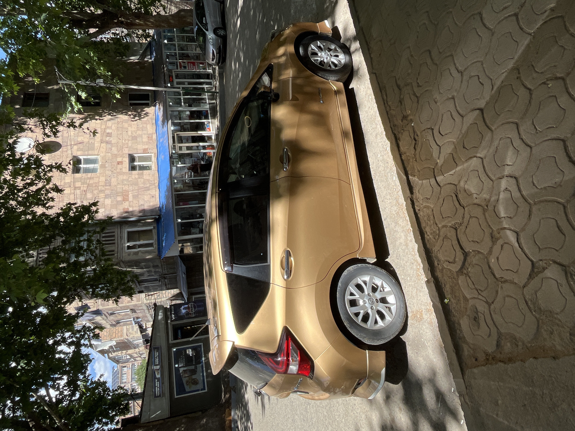 This media file captures a street scene with a golden car as the central object. The car is parked on the right side of a sidewalk with a patterned pavement. To the left of the car, a building with a stone facade is visible. This building appears to house shops on its ground floor, indicated by storefronts with glass windows displaying various items. A blue awning is stretched over one of these storefronts. The scene is framed by the branches of a large tree on the far left, casting dappled shadows on the car and the pavement. The overall lighting suggests it is daytime, likely late morning or early afternoon, due to the strong shadows.
There are no people clearly visible in the image, though the storefronts suggest a public area. The car is a hatchback, with its rear and right side prominently displayed. The car's design includes modern taillights and alloy wheels. The building shows architectural details typical of older urban structures, with multiple windows on its upper floors. The tree's foliage is lush and green, suggesting a temperate climate during a warmer season.
There is some visible text on signs within the storefronts, but it is not legible enough to be deciphered. The overall impression is a snapshot of a quiet urban street on a sunny day.