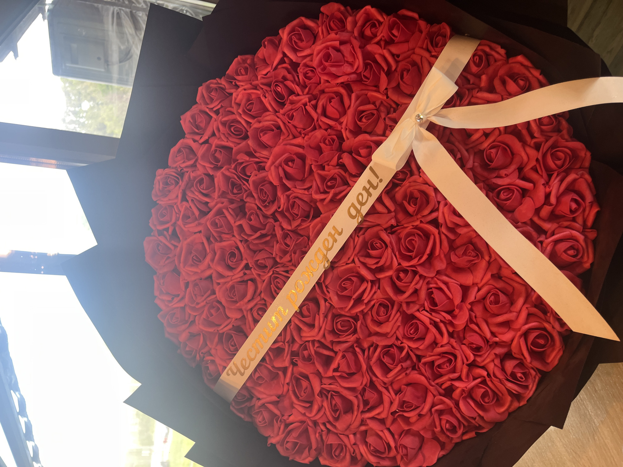 A close-up, slightly angled shot shows a bouquet of numerous red roses tightly arranged in a circular formation. The roses appear artificial with a matte texture. The bouquet is wrapped in dark grey paper. A wide, off-white ribbon is tied around the bouquet with a decorative rhinestone detail at the knot. A second ribbon with text in Cyrillic, "Честит рожден ден!", runs diagonally across the roses. The bouquet is partially in sunlight near a window. The location is in the city of Plovdiv, Bulgaria.