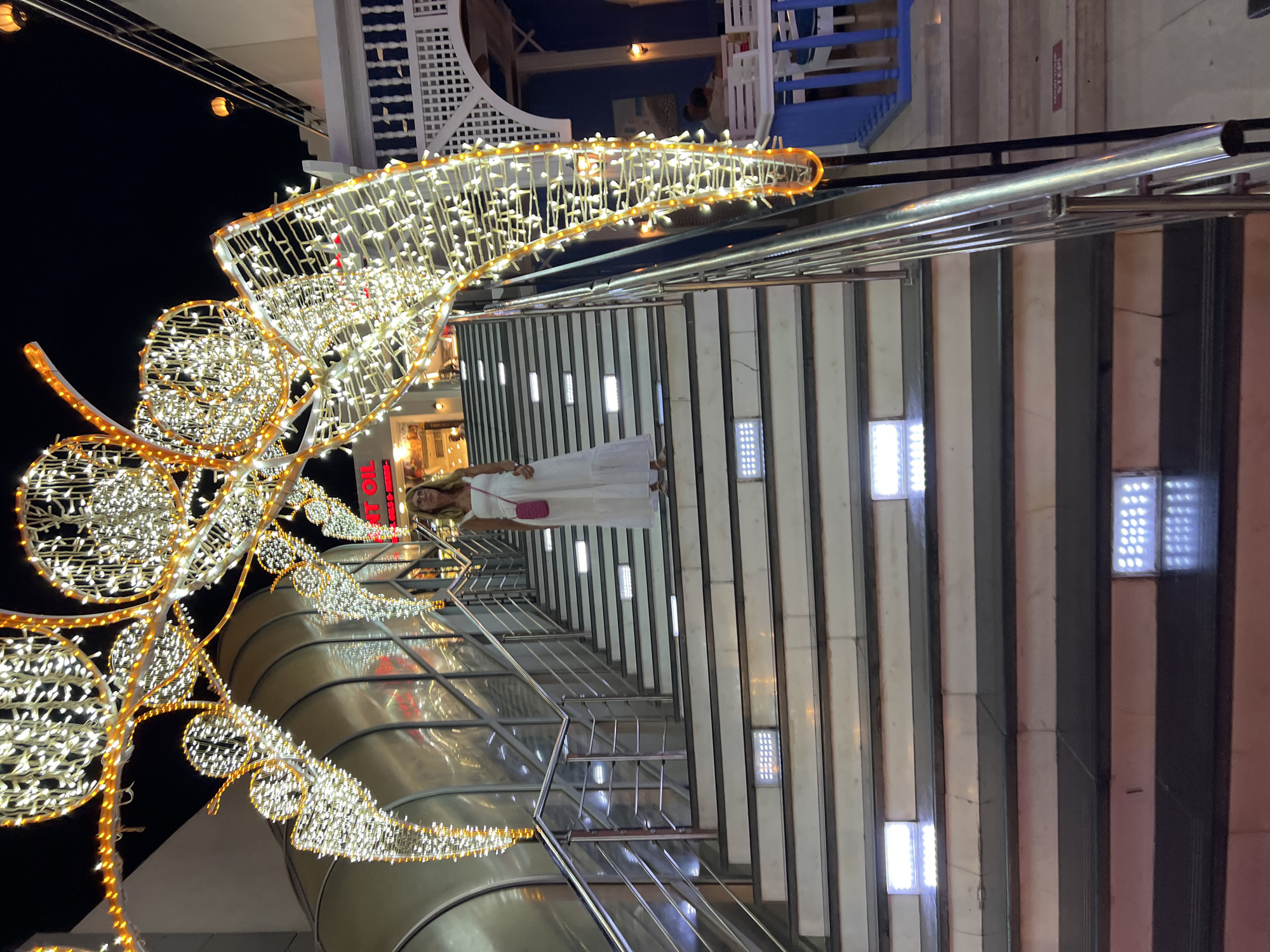 A full-shot, eye-level, vertical night-time photograph captures a woman in a white dress standing on a wide, outdoor staircase. The staircase is illuminated by bright, white LED lights embedded in each step, and bordered by a silver railing. Above the staircase, an ornate archway constructed of string lights, resembling a butterfly or floral motif, is festooned with bright white and amber lights. The woman, appearing to be in her late 30s, wears a sleeveless white maxi dress and carries a small, bright pink crossbody bag. Her blonde hair is pulled back, and she is looking towards the camera with a slight smile. The background reveals shops with red and white signage, one reading "AT OIL", and a dark night sky. In the upper right quadrant, a white, lattice-style structure with blue accents suggests an outdoor seating area, possibly a restaurant or cafe, with tables and chairs visible. The scene is located in Sharm El Sheikh, Egypt.